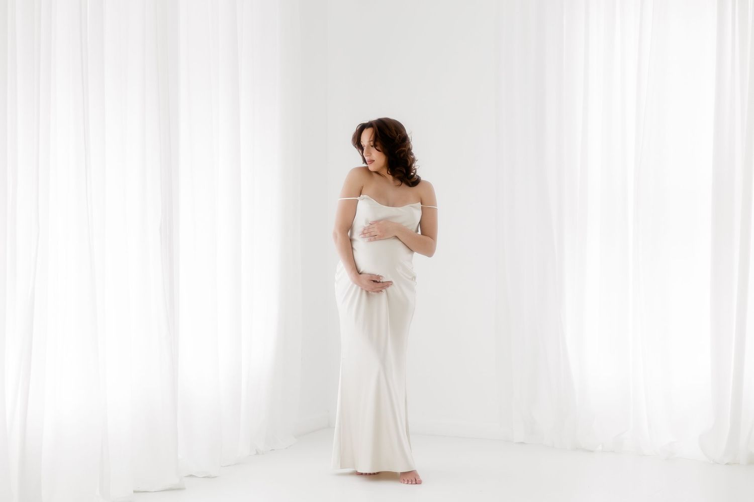 A bride in a white wedding dress poses elegantly against sheer white curtains in a bright, airy room.