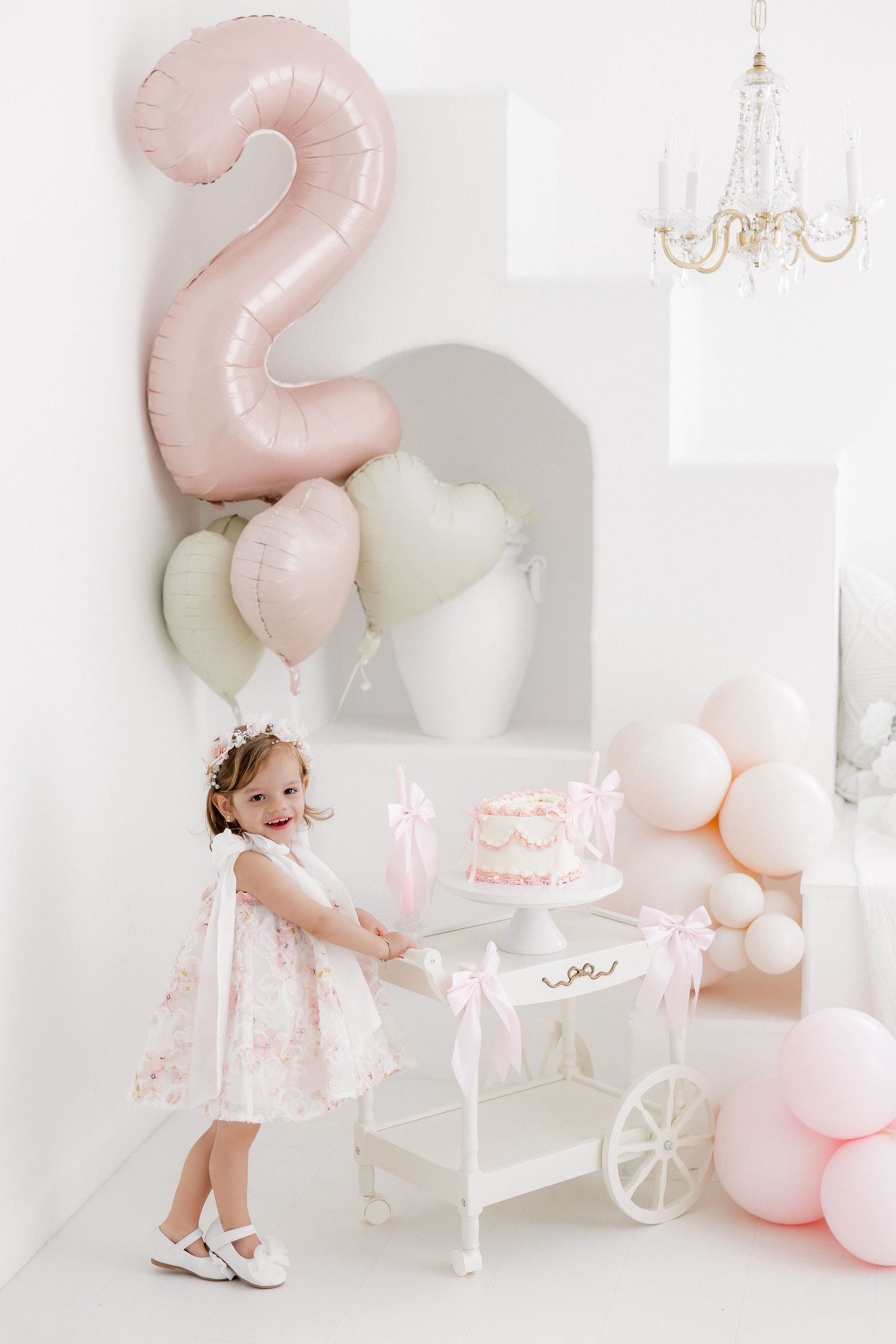 Pink and white birthday party setup with balloons, decorative cart, chandelier and elegant decor in a bright studio setting.