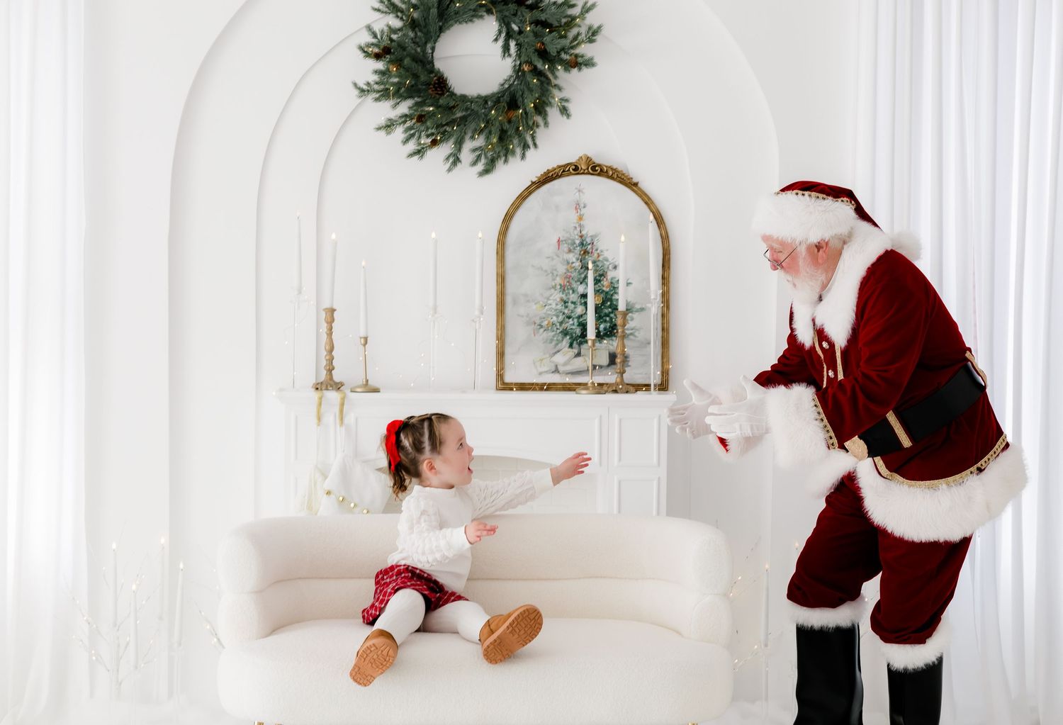 A toddler in red and white sits on a cream sofa reaching toward Santa Claus in a bright white holiday room.