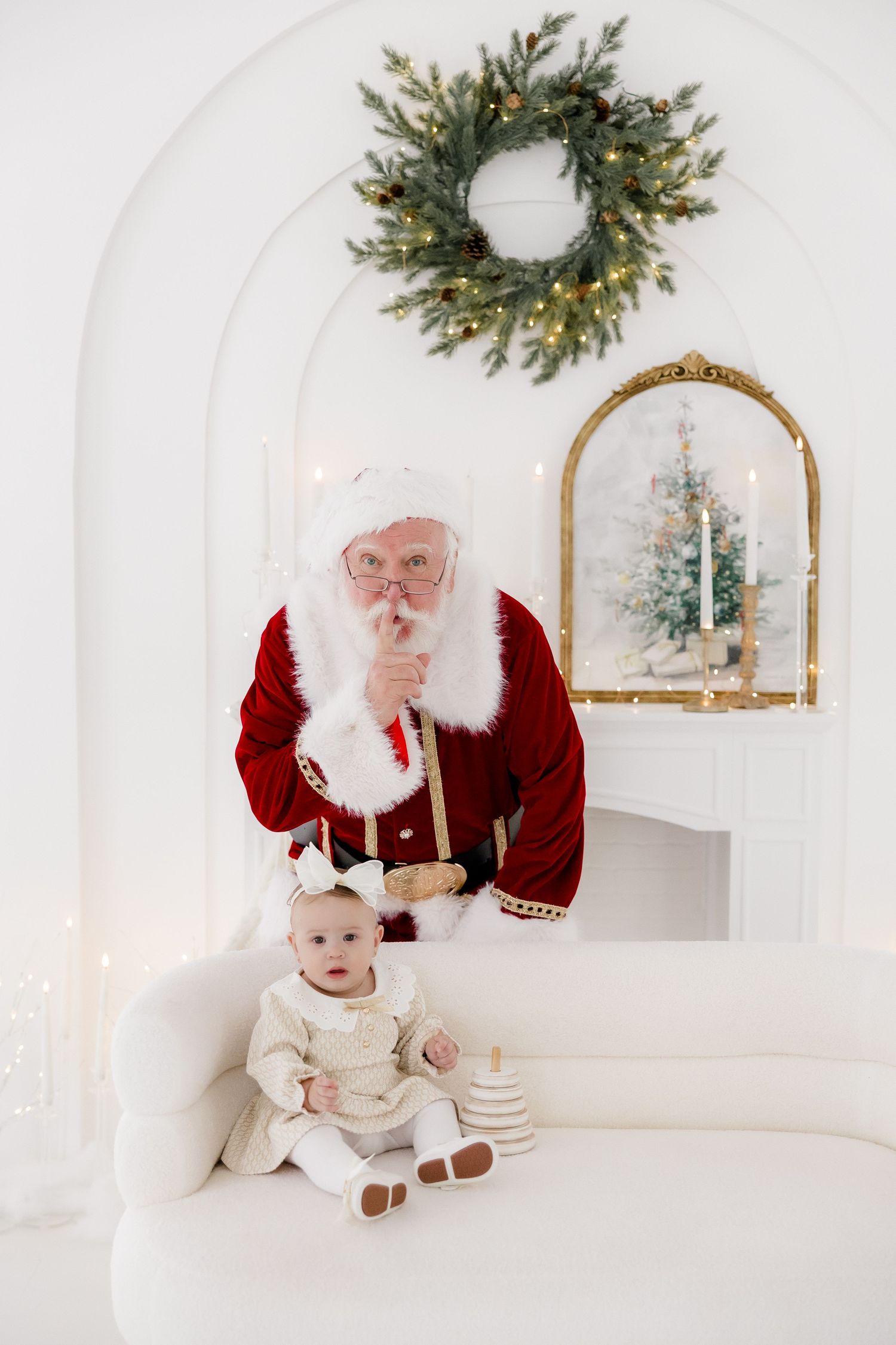 Santa Claus poses with a baby in white outfit on a sofa, Christmas wreath and fireplace in background.