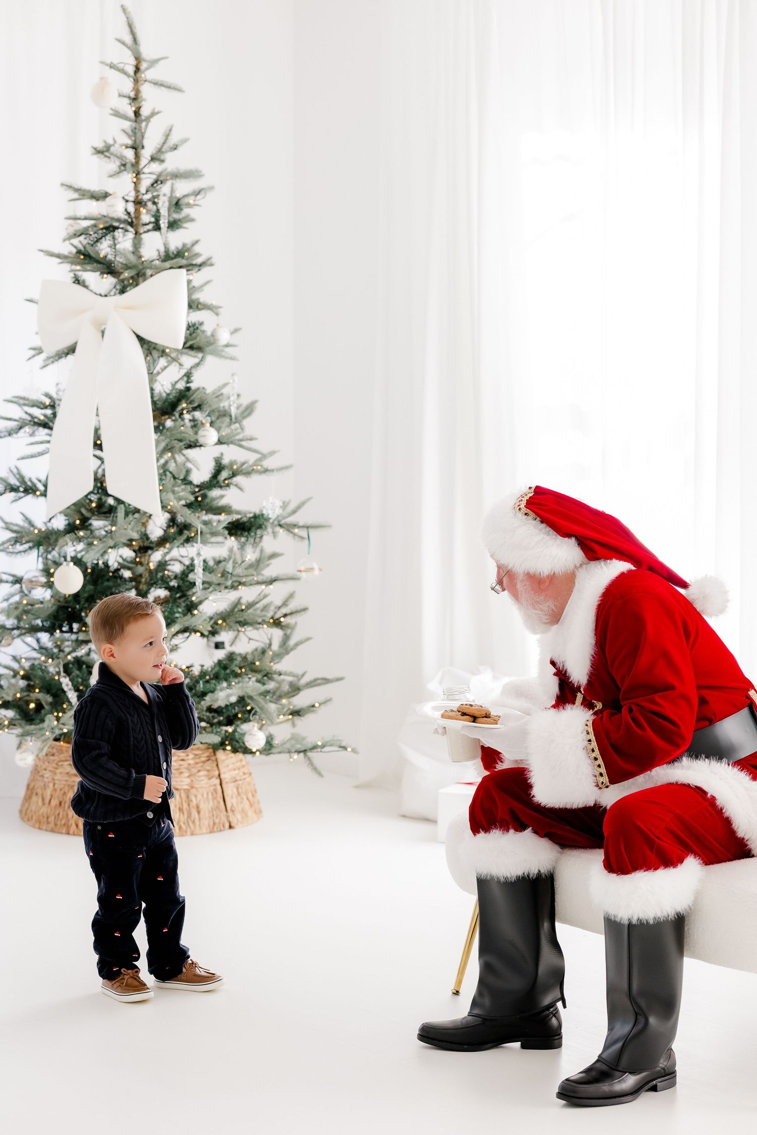 A young boy meets Santa Claus beside a decorated white Christmas tree in a bright, festive room.