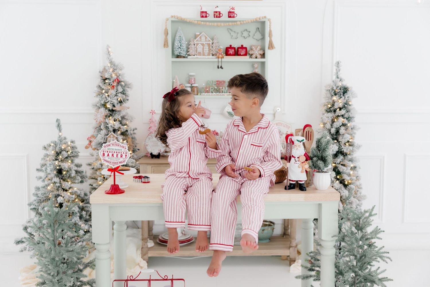 Two children in matching red striped Christmas pajamas sit together on a white bench surrounded by festive holiday decor.