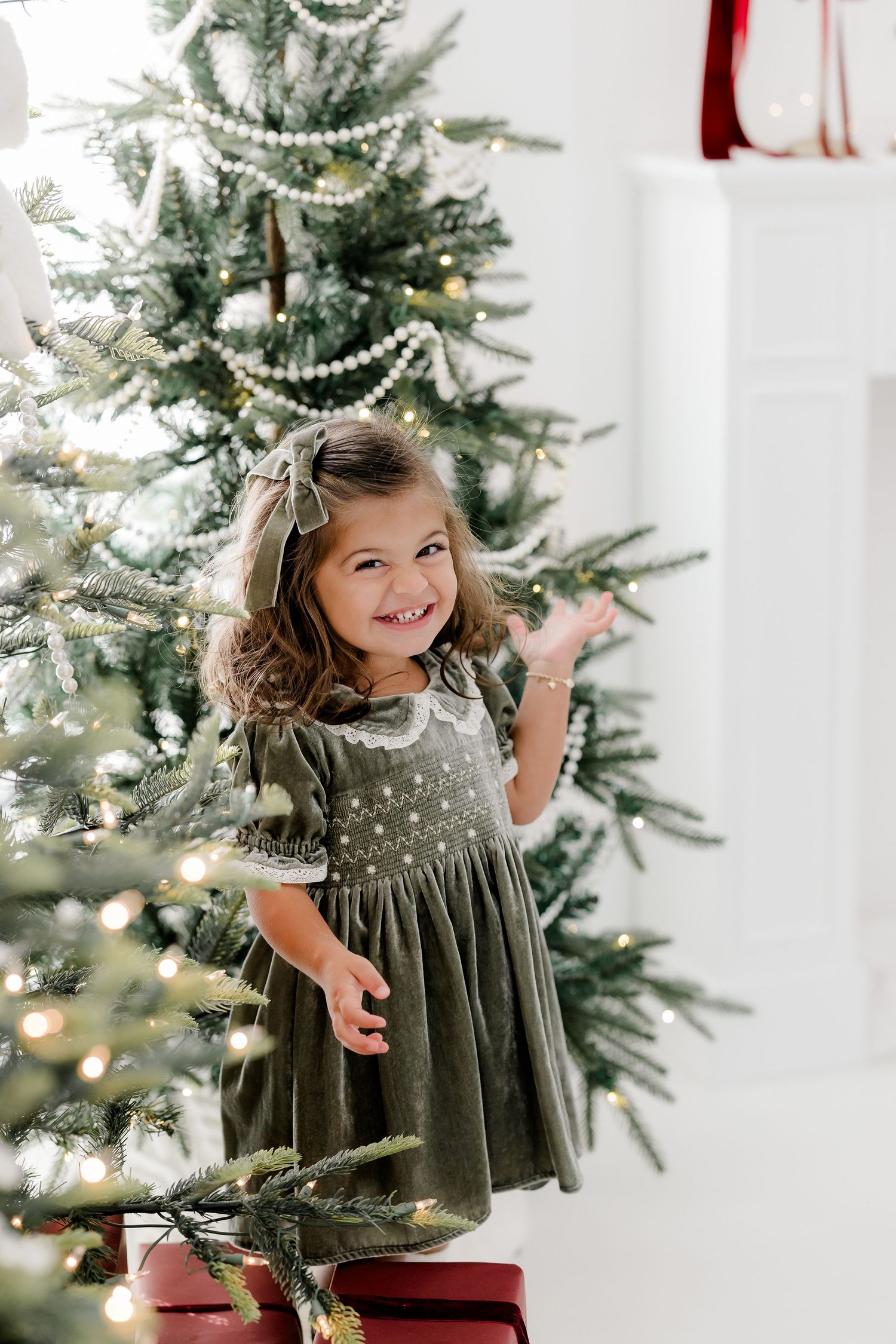 Young girl in green velvet dress smiles beside decorated Christmas tree with pearl garland and twinkling lights.