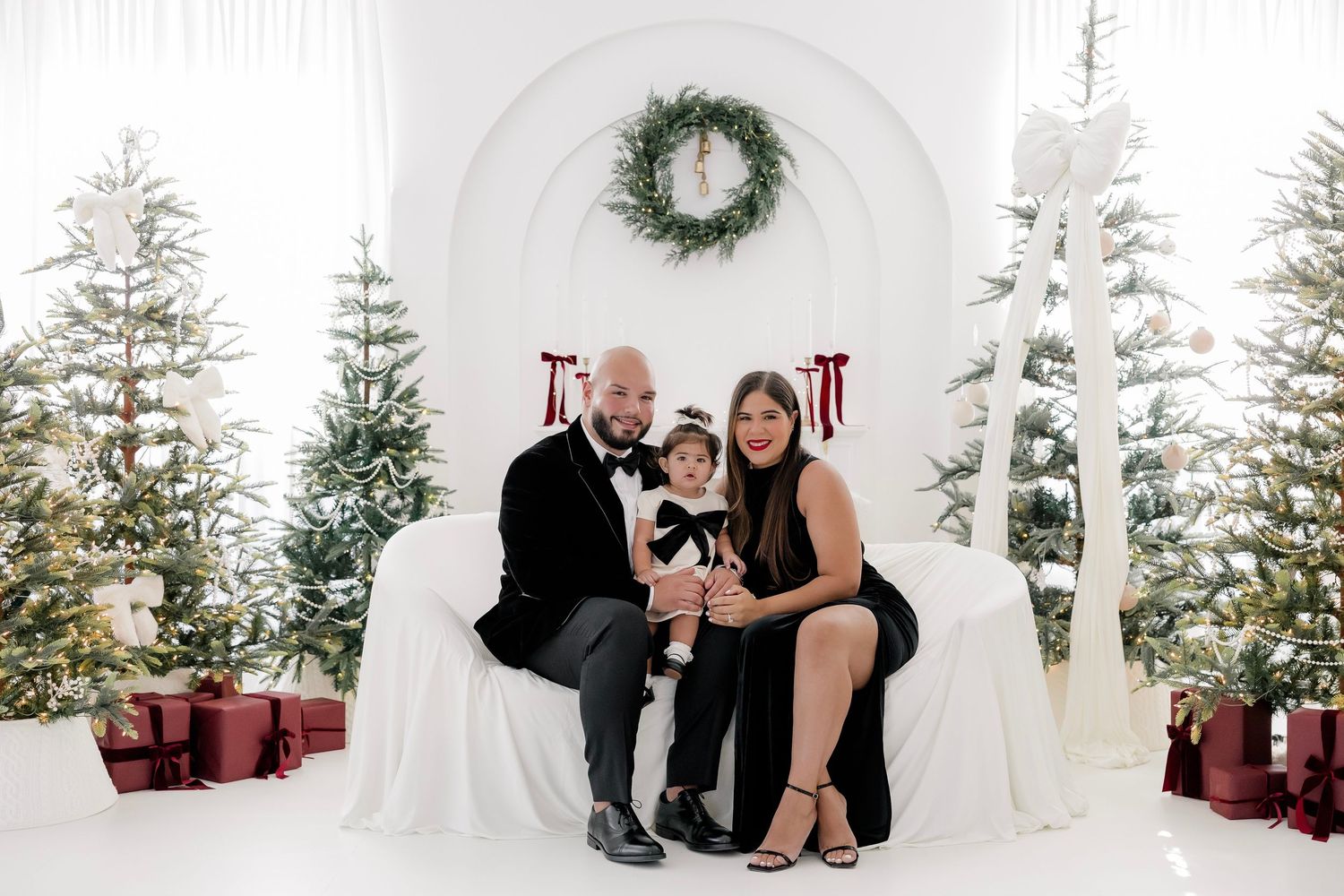 Family of three posing for a Christmas portrait with decorated trees, gifts, and a wreath in a white studio setting.