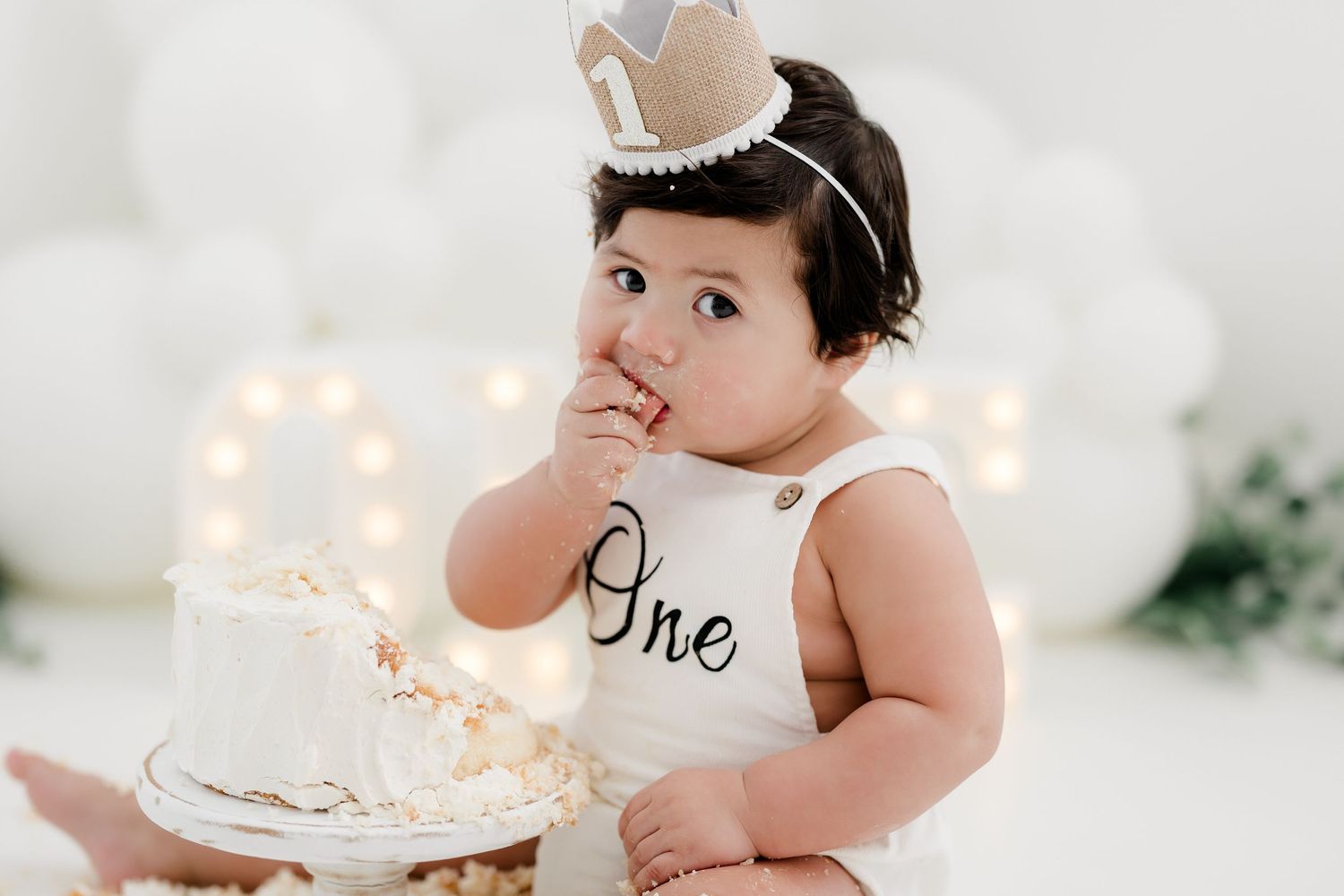 Baby in white 'One' onesie tastes birthday cake while wearing small crown.