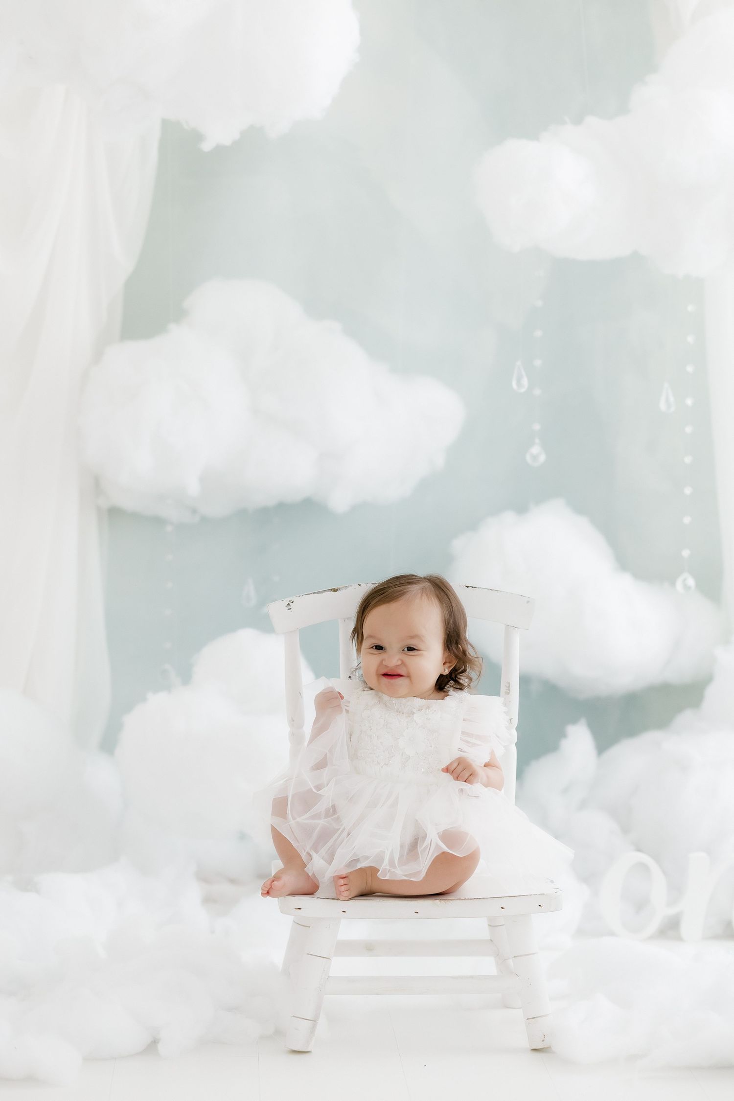 Smiling baby girl in white tulle dress sits on white chair in dreamy cloud-themed photo studio.