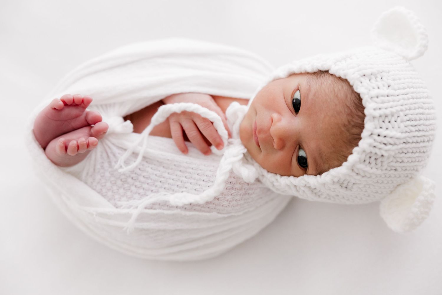 A baby wrapped in white fabric lying on soft linens in a minimalist setting.