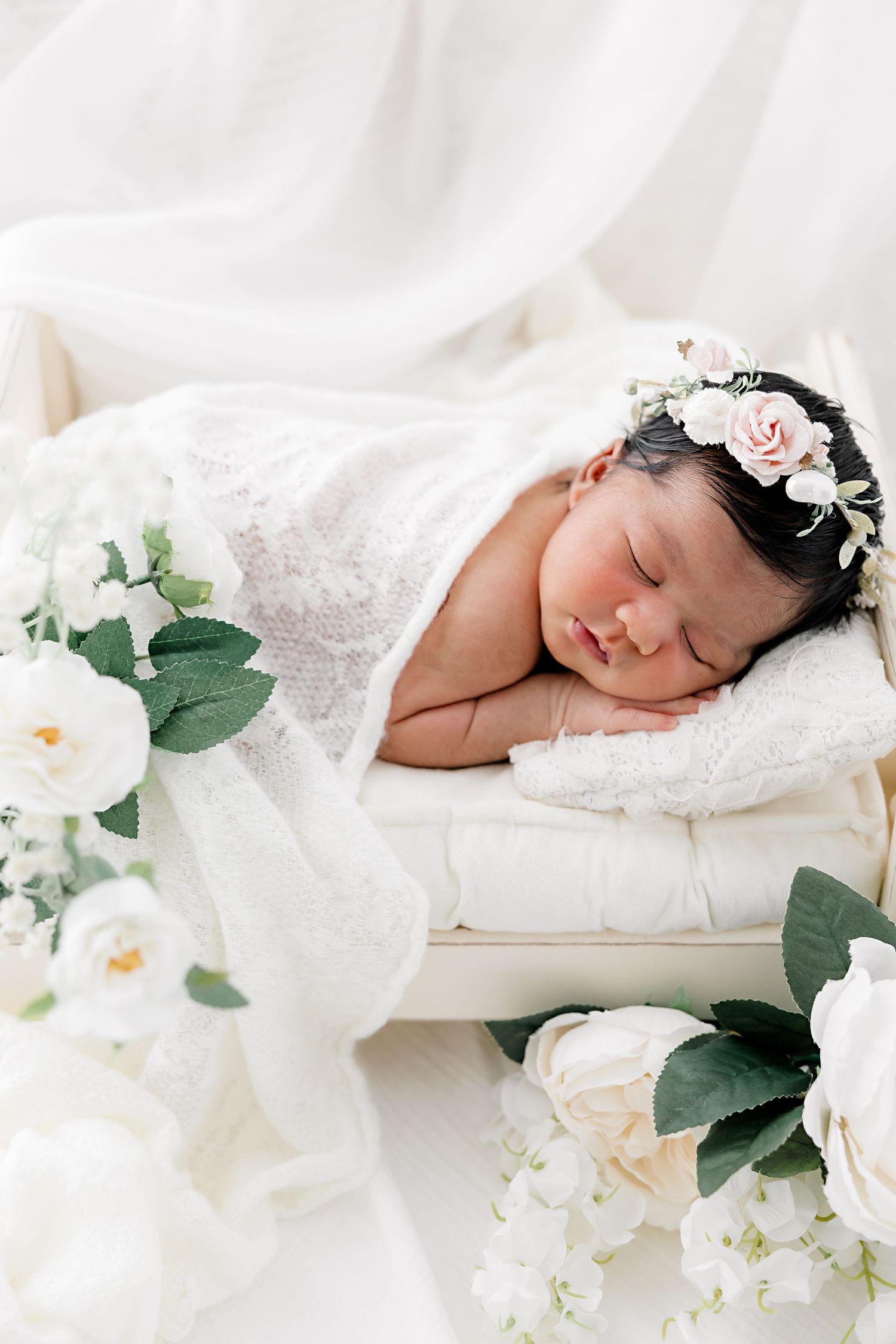 Peaceful newborn sleeping among white florals and delicate lace in ethereal setting.