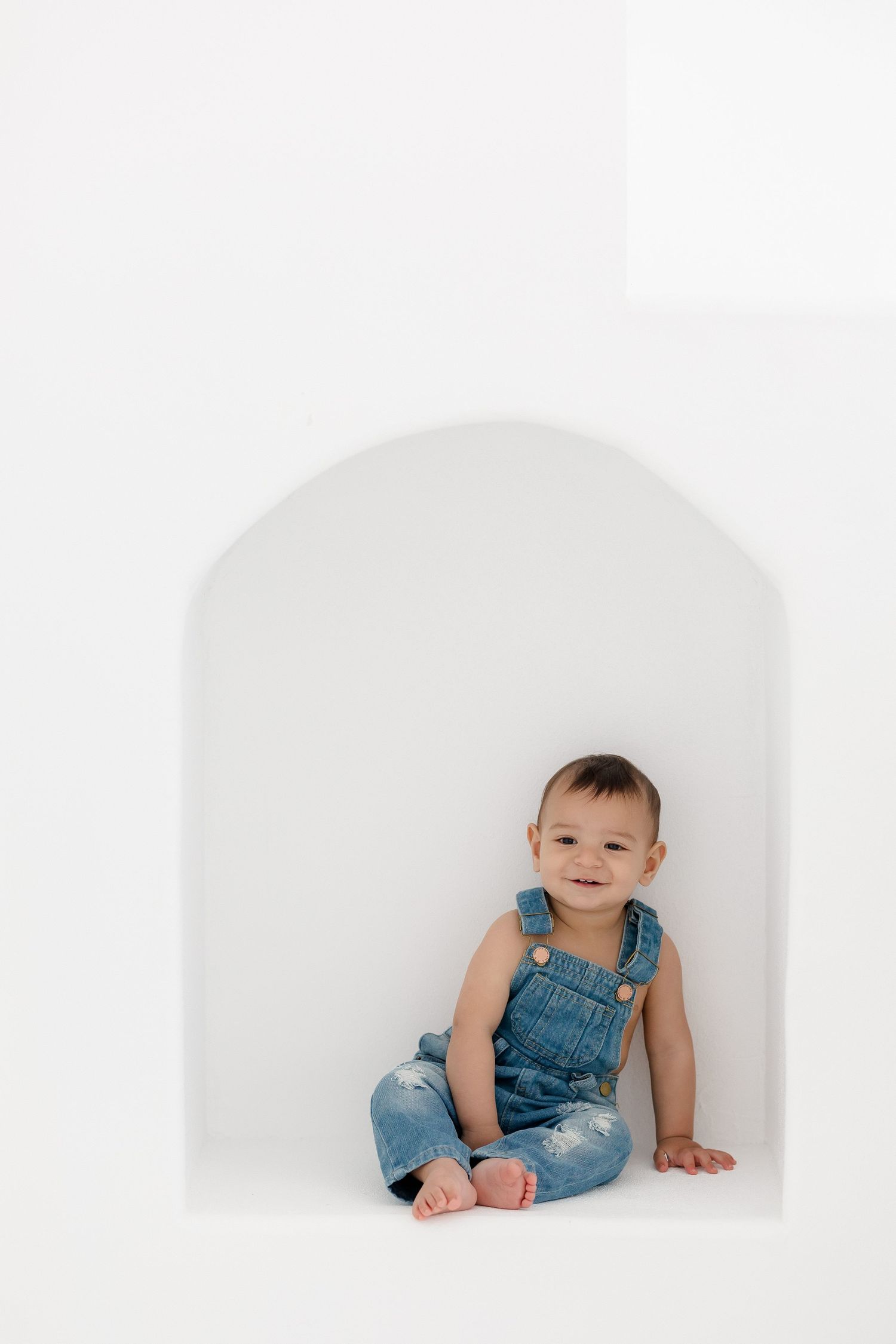 Baby wearing blue denim overalls sitting in white alcove setting.