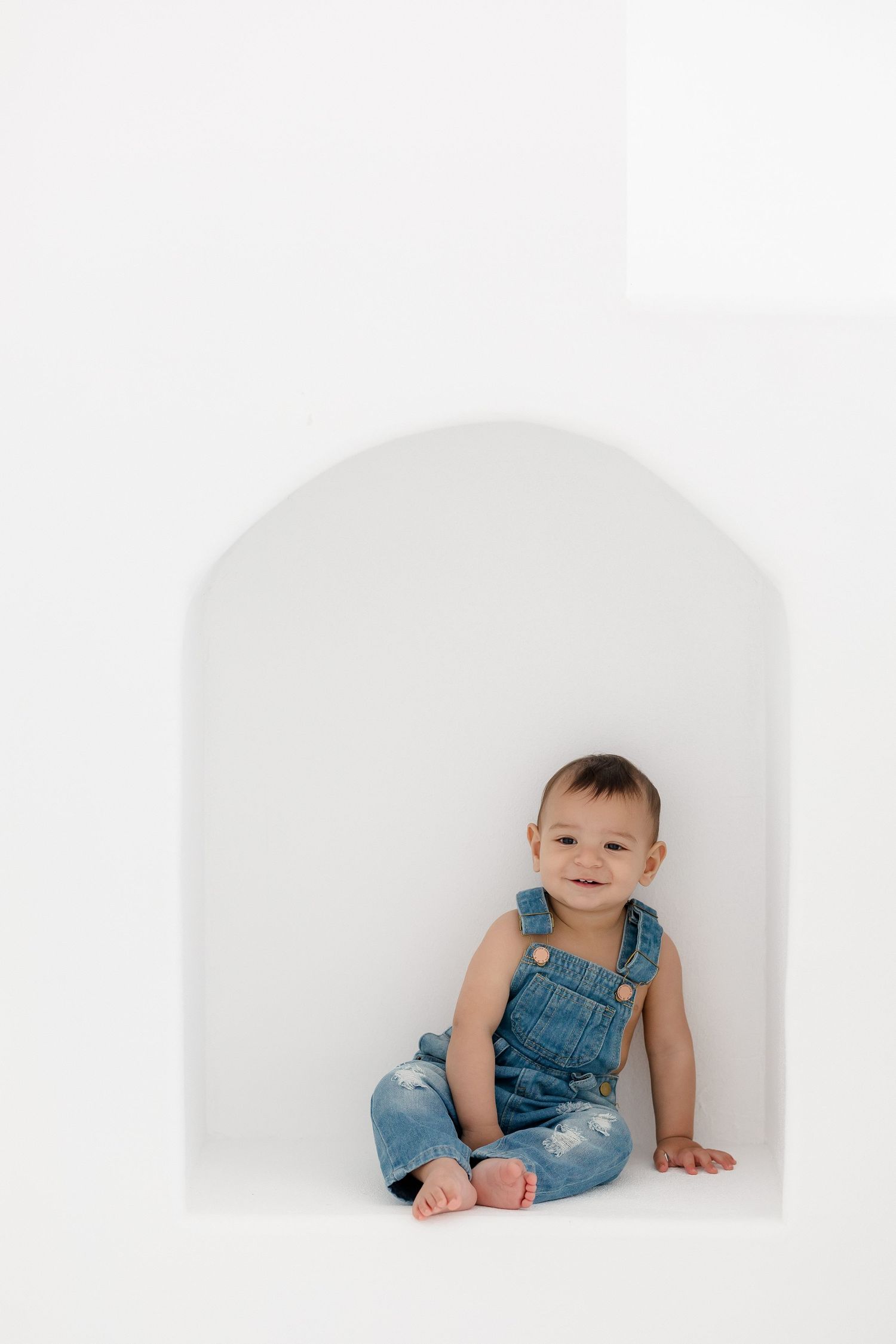Baby wearing denim overalls sits and crawls on white background in studio portrait series.