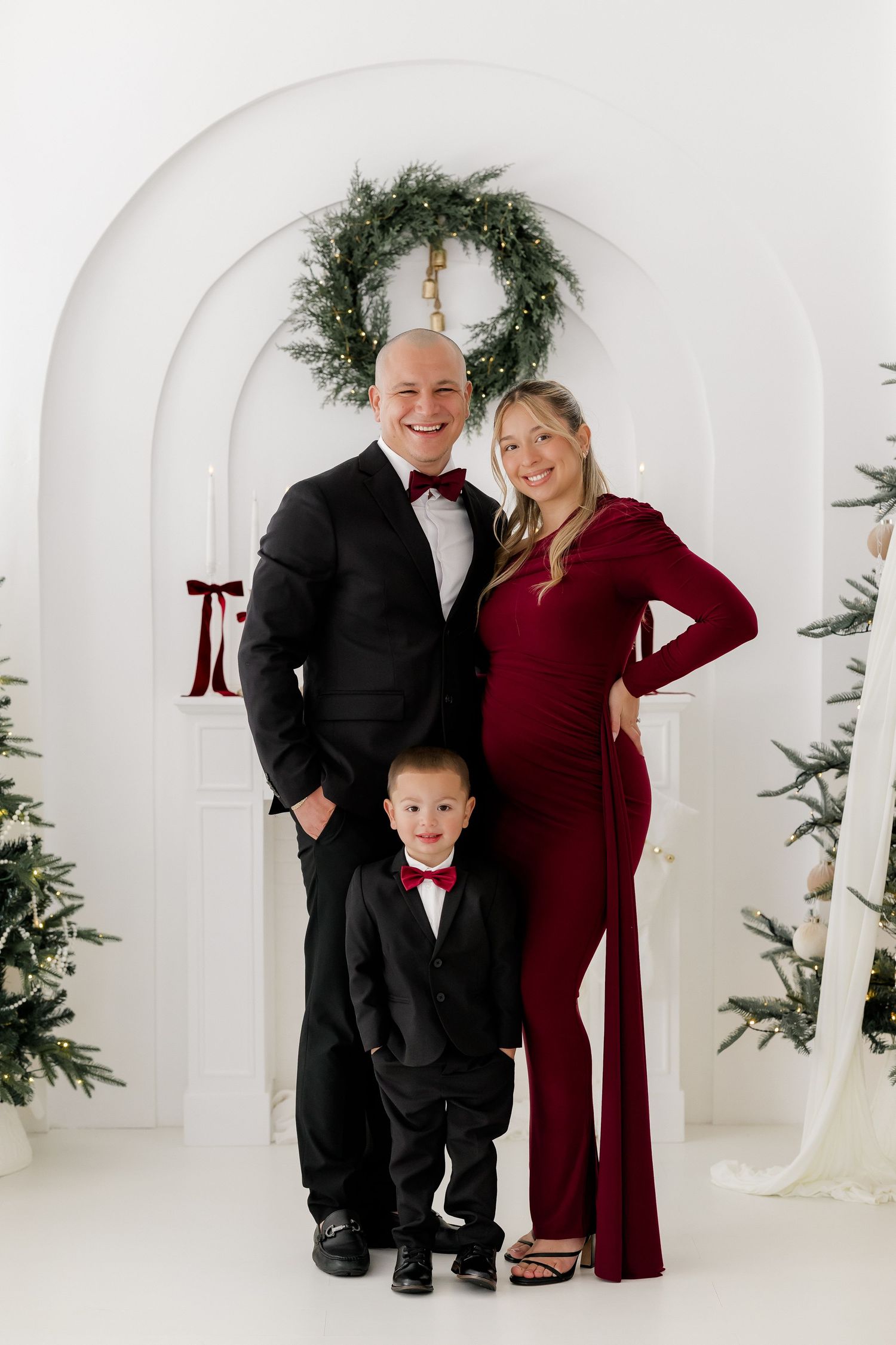 Family Christmas portrait with pregnant mother in burgundy dress, father in tuxedo, and young son in matching suit.