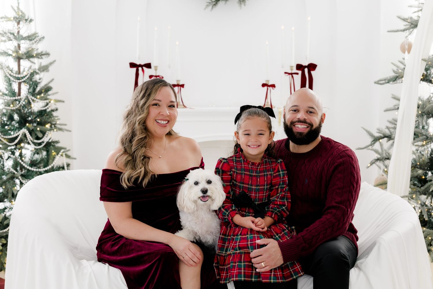 Family of three with a white dog poses in festive holiday attire beside decorated Christmas trees.