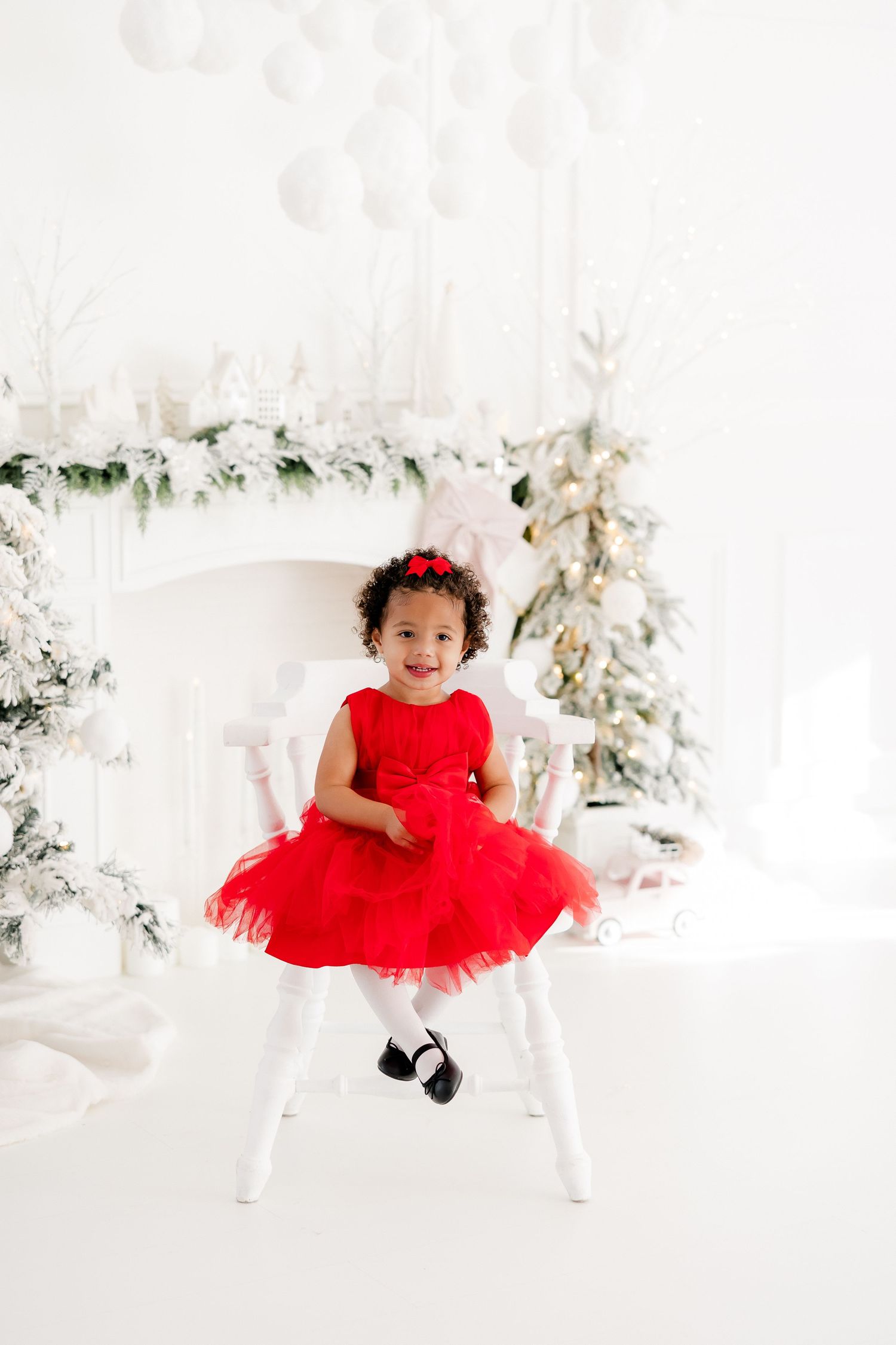 A young child in a festive red tulle dress sits on a white chair surrounded by holiday decorations.