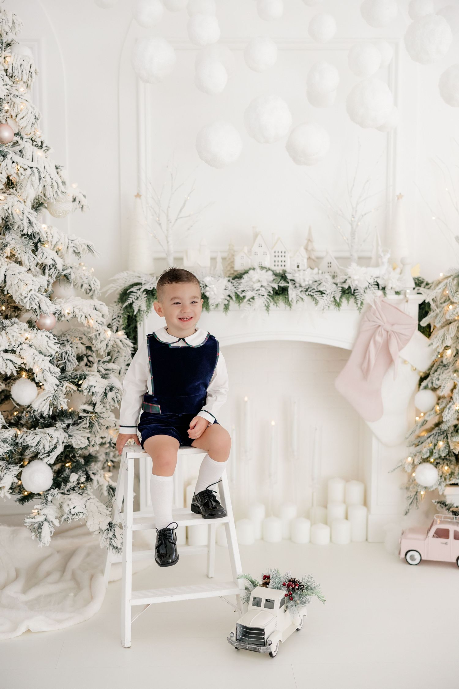 Child in navy outfit sitting on white stool beside Christmas decor and toy cars.