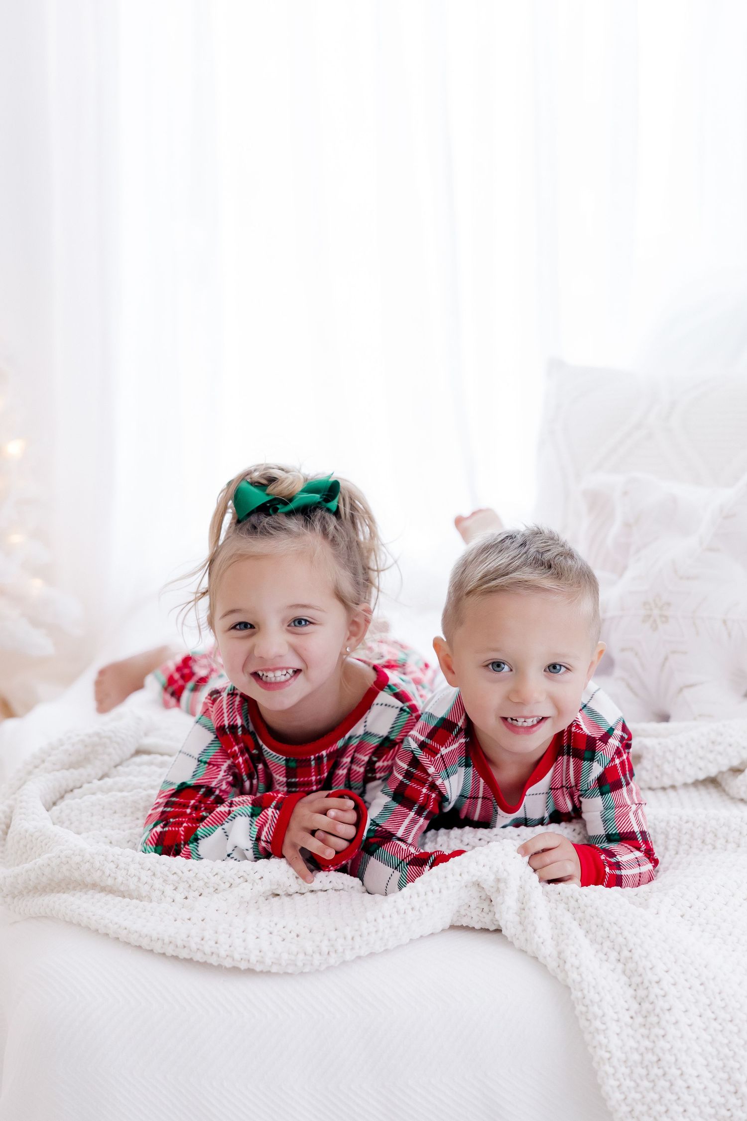 Two children in matching plaid pajamas lying on white blanket during Christmas.