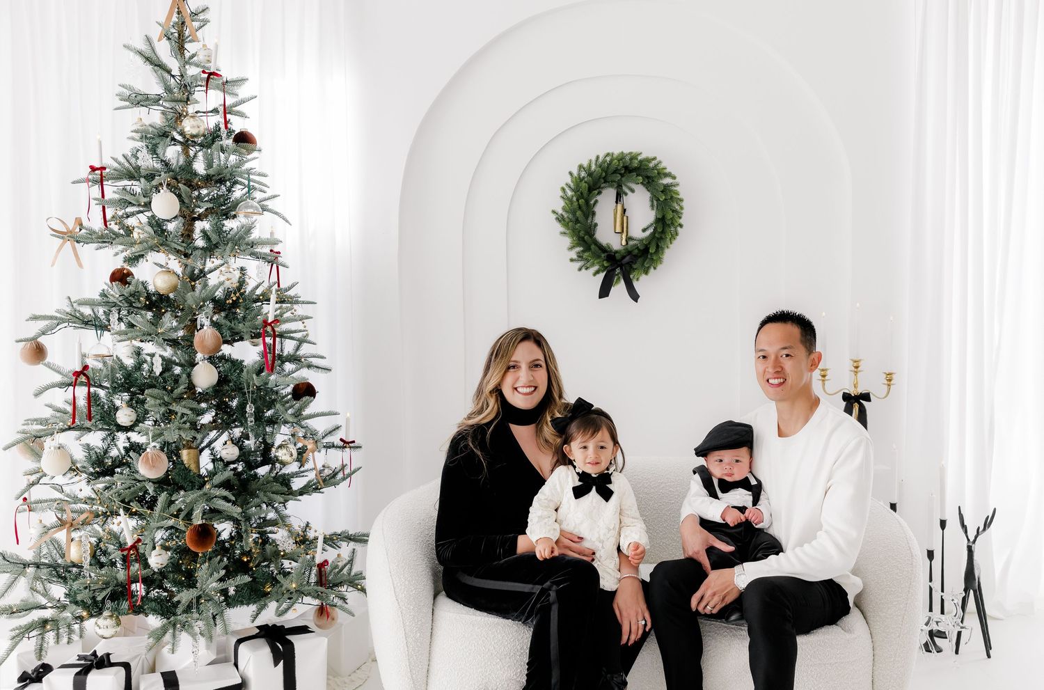 Family poses together in white studio with decorated Christmas trees and festive wreaths.