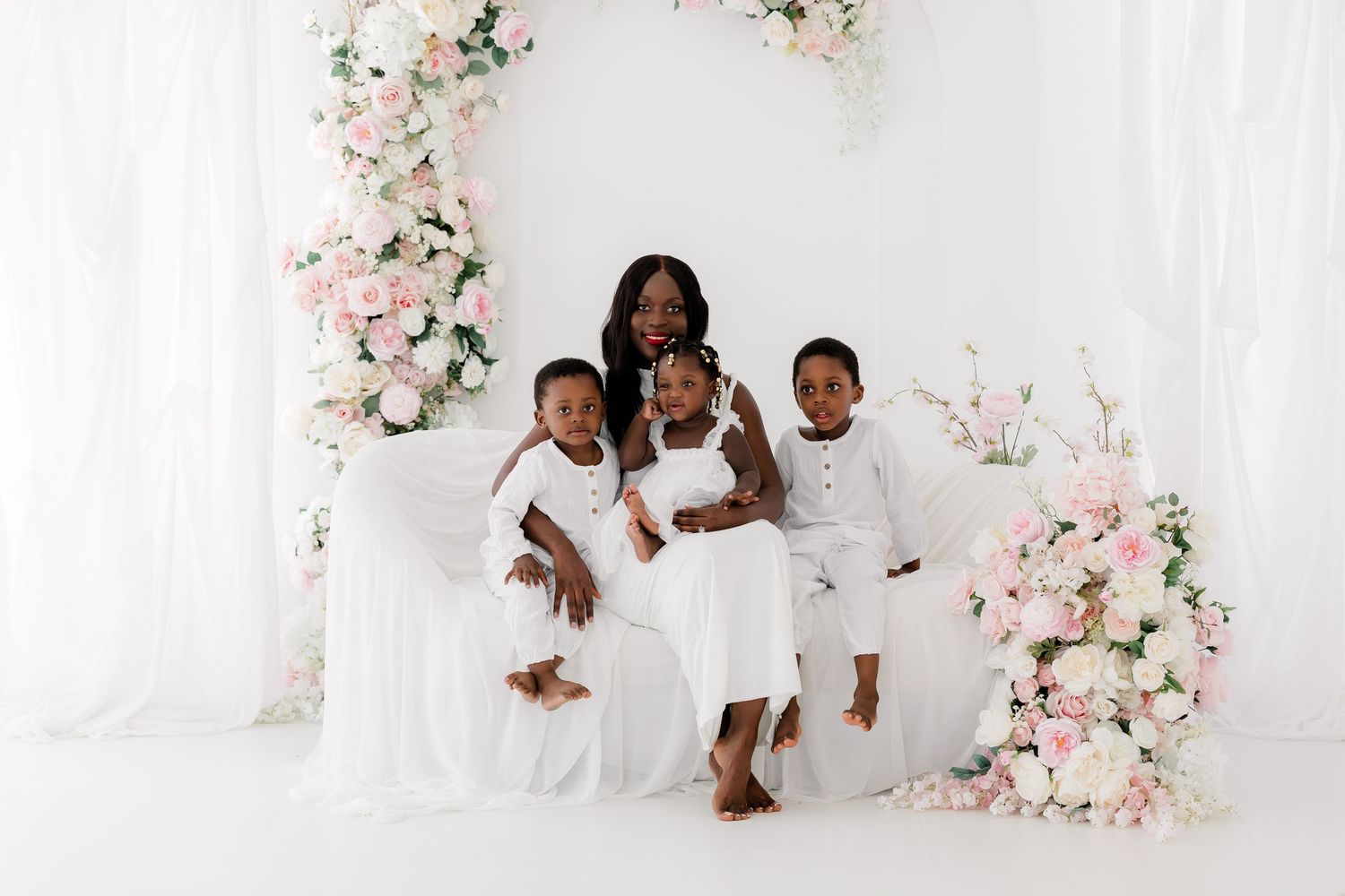 Beautiful family portrait in all white outfits against a white backdrop with pink floral decorations.