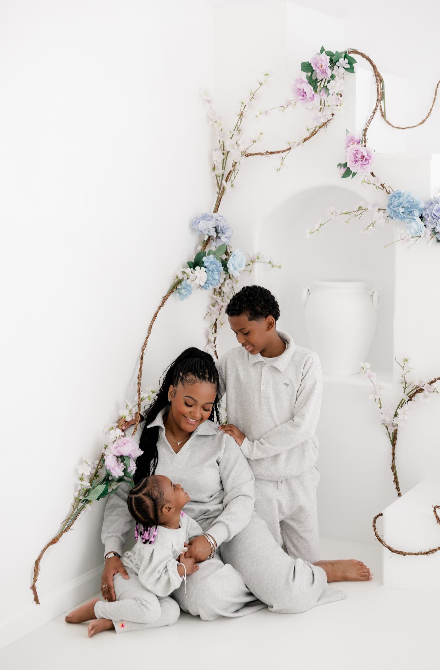 Family wearing matching white outfits poses together against white wall decorated with delicate floral vines and blossoms.