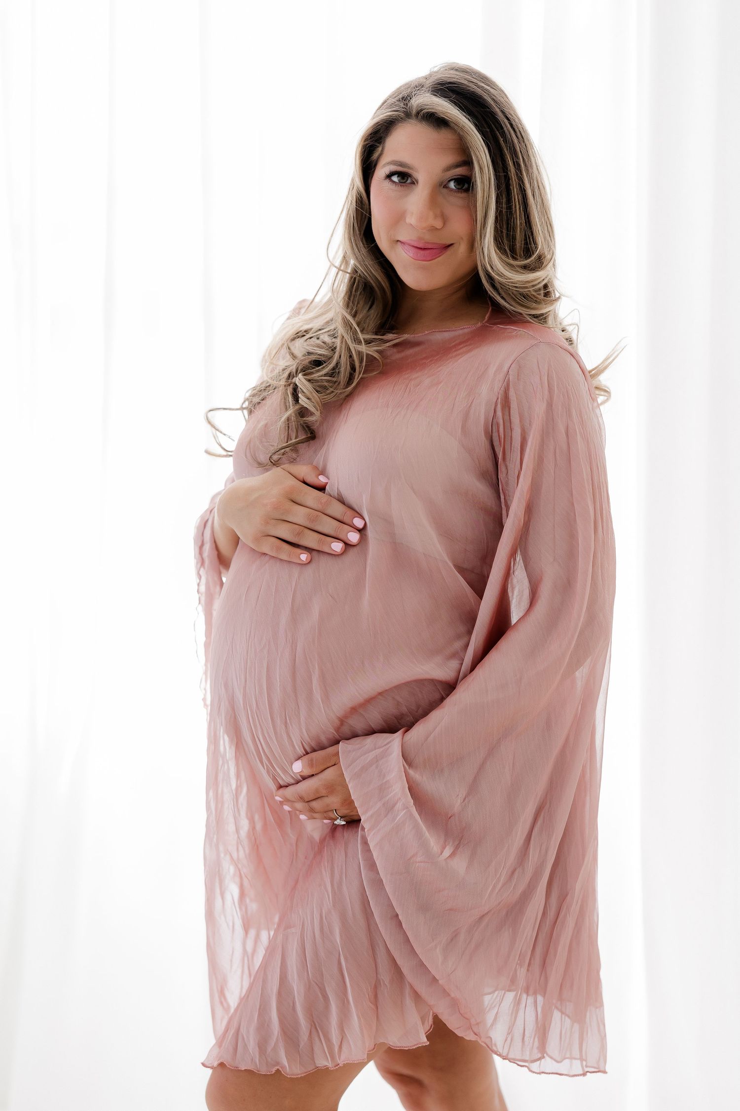 Maternity photo series showing a woman in a flowing pink dress against a white backdrop.