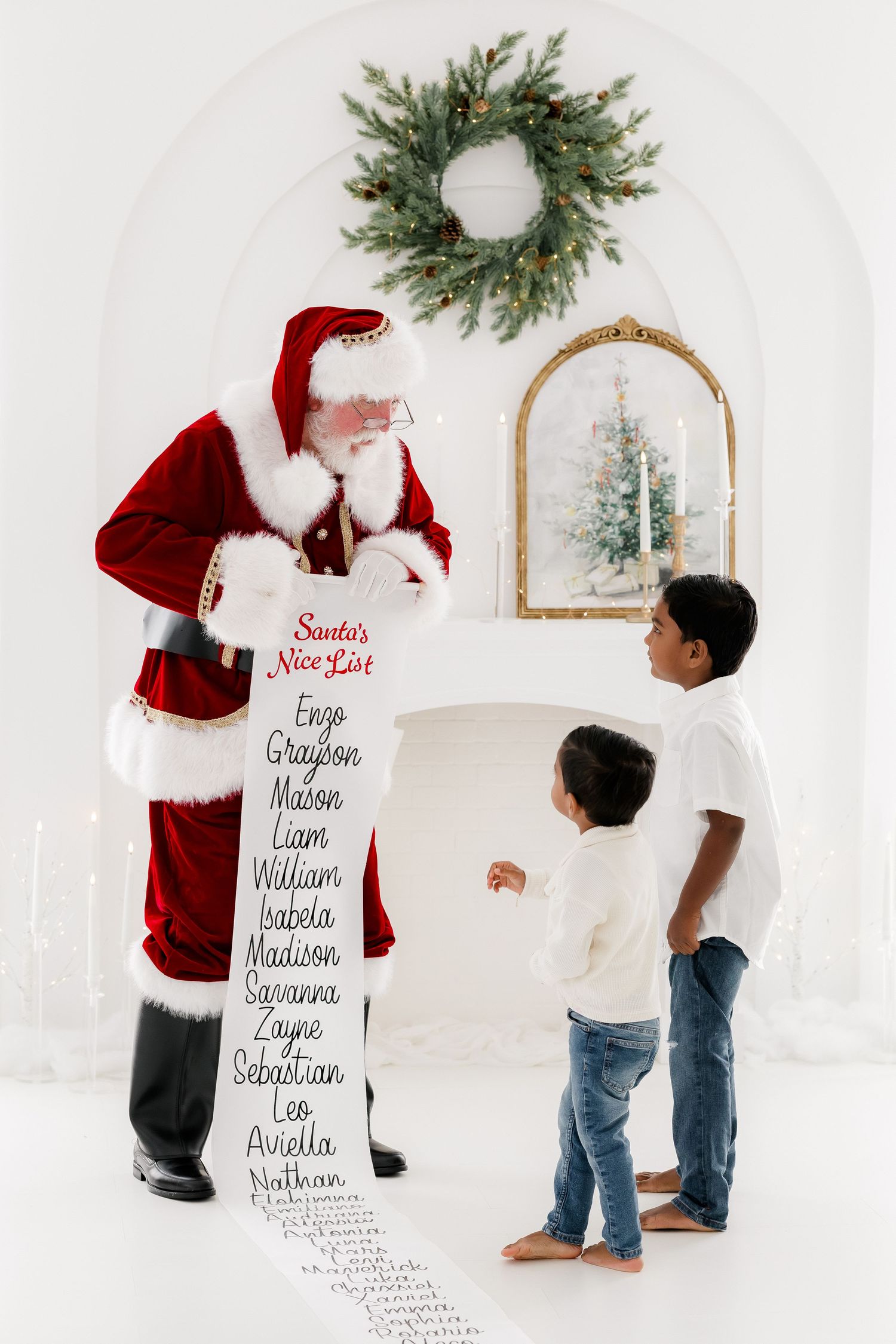 Santa Claus shows two young boys his Nice List scroll in a white Christmas-decorated room with wreath and fireplace.