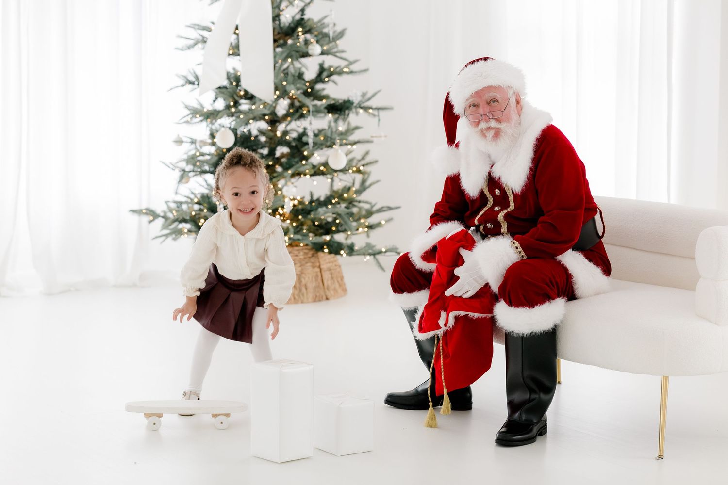 A young girl plays near gift boxes beside Santa Claus sitting in a white room with a decorated Christmas tree.