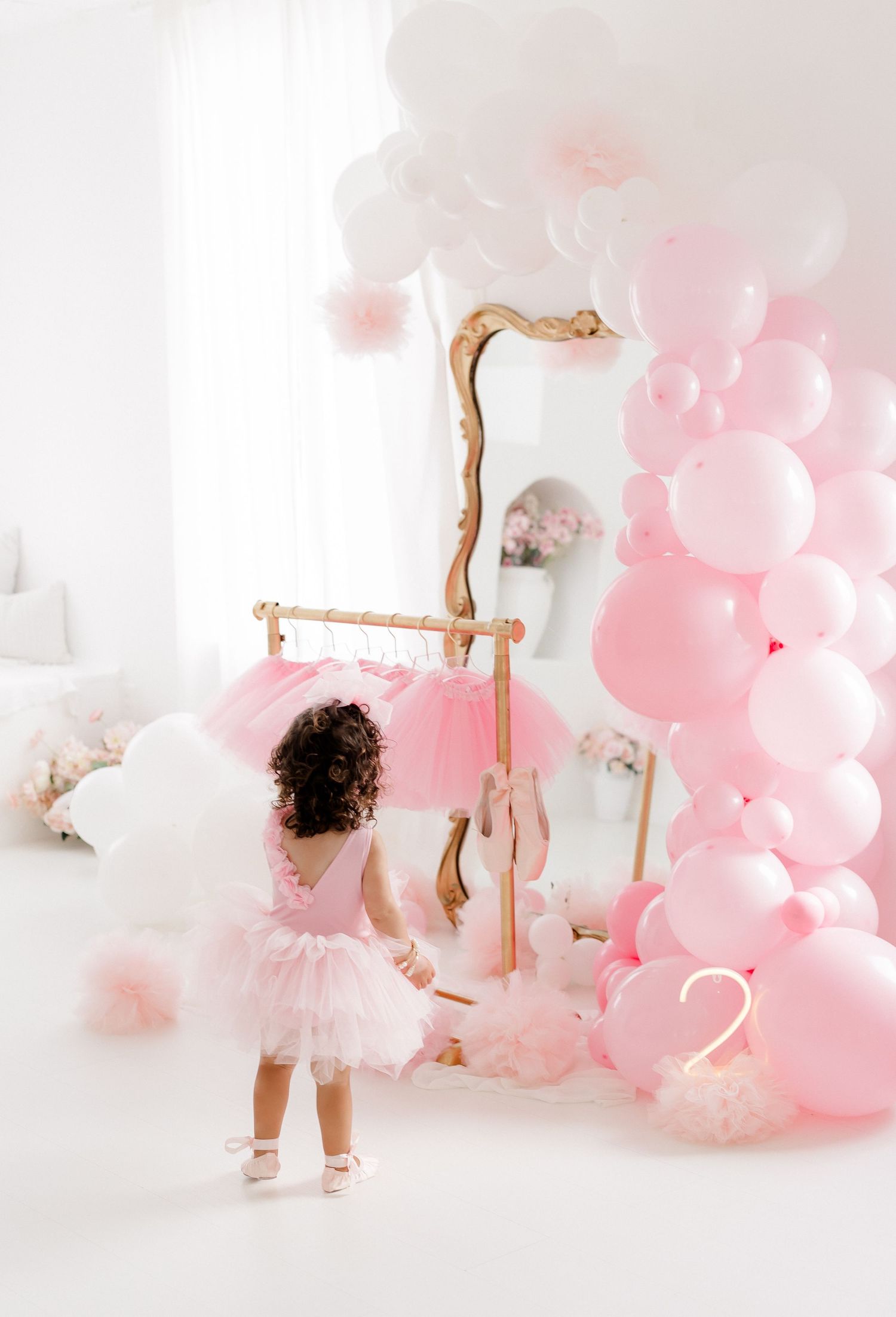 A child in a white tutu dress plays among pink and white balloons in a dreamy studio setting.