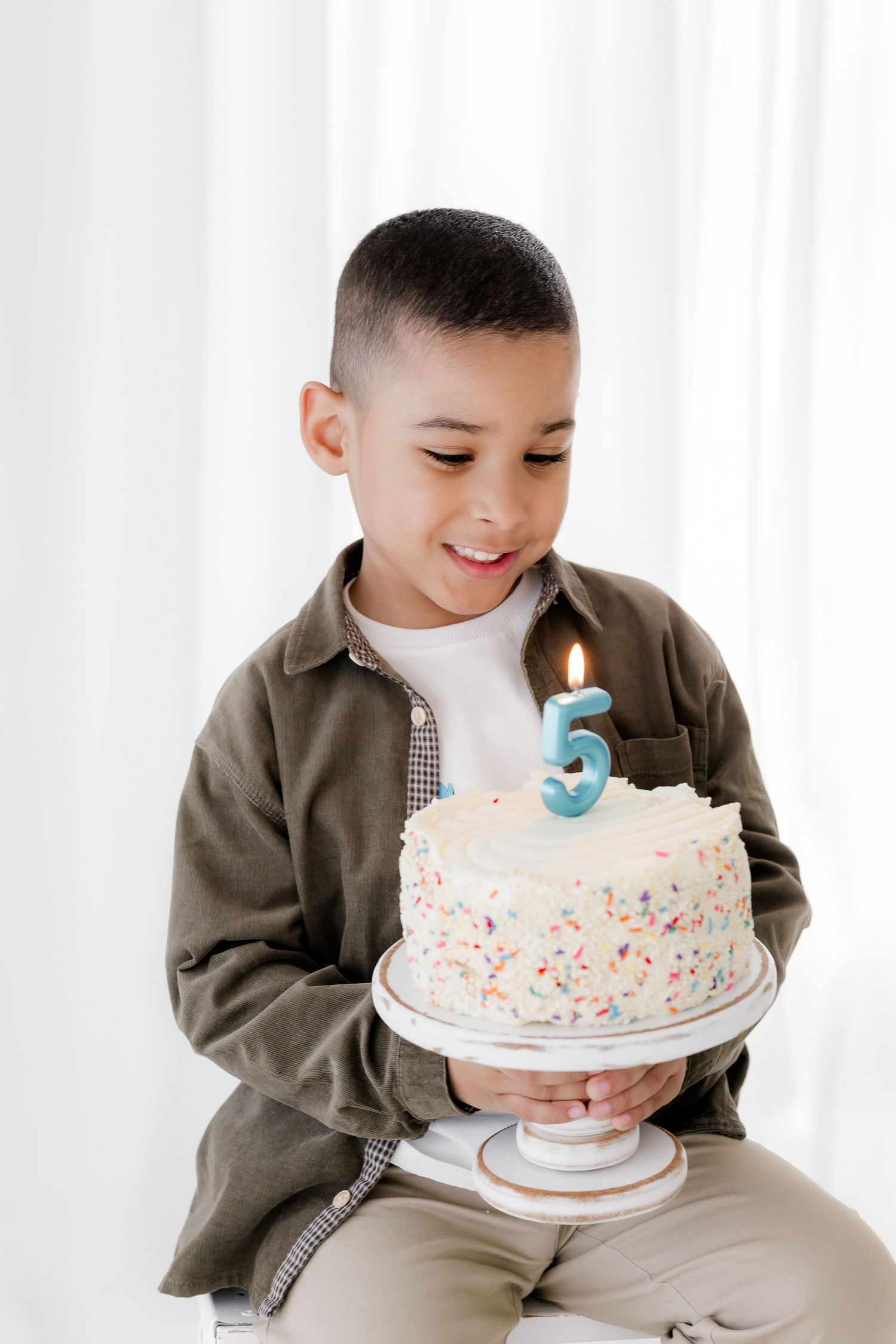 Child in brown jacket sits with white birthday cake decorated with sprinkles and blue number 3 candle on white background.