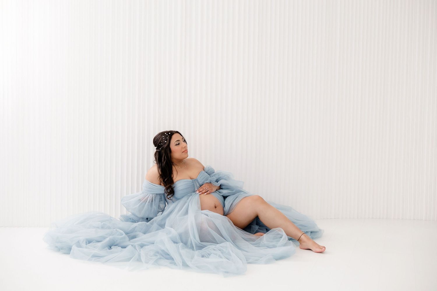 Dreamy maternity pose on white backdrop with flowing powder blue fabric.