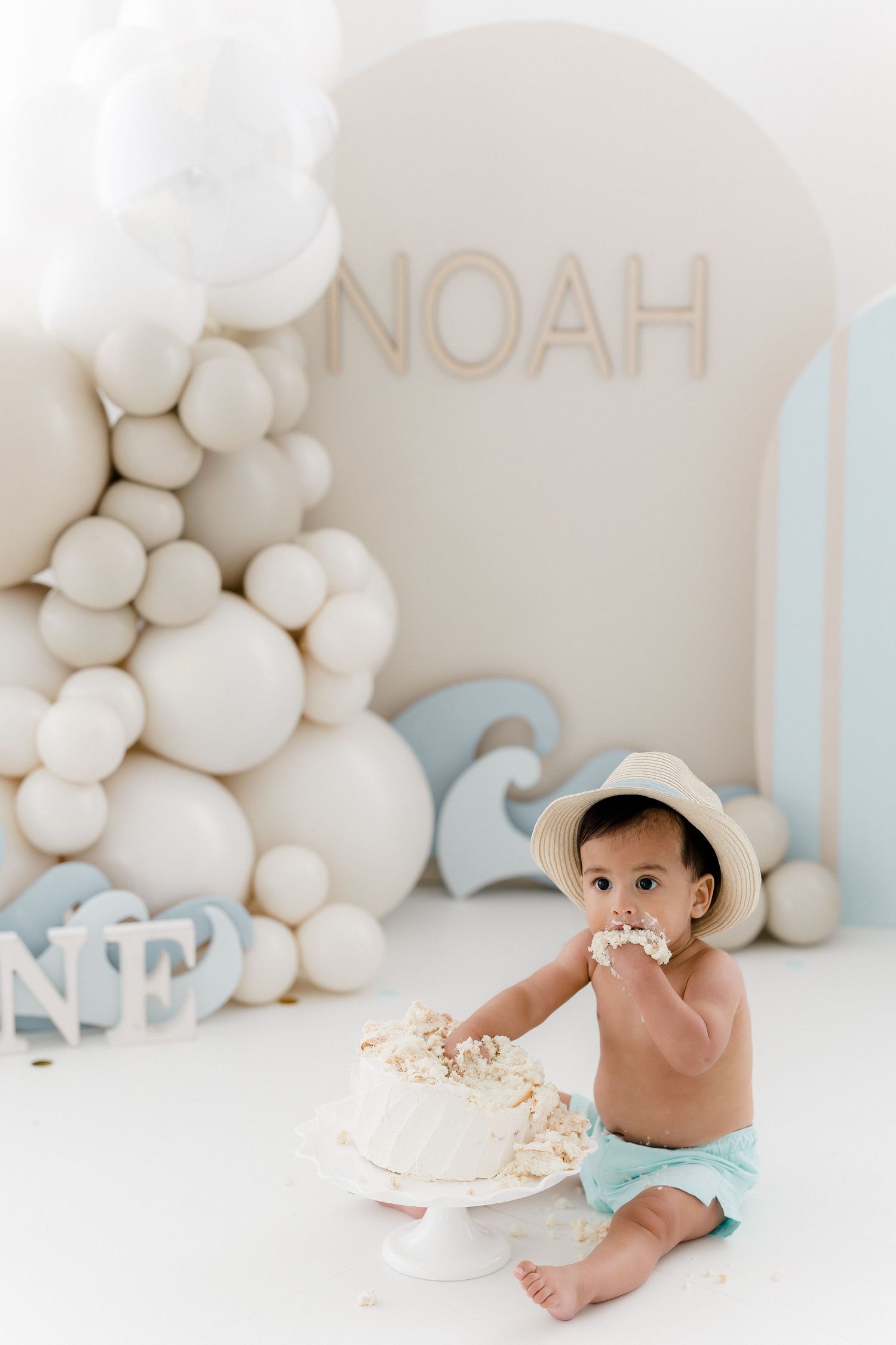 Baby in white diaper sitting against soft white balloon backdrop with blue accents during cake smash photo session.