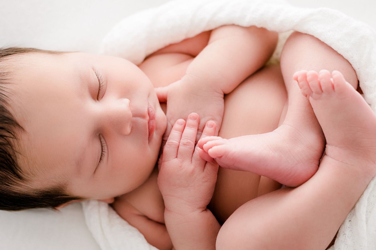 A newborn baby's tiny feet and hands are gently curled while sleeping peacefully on white bedding.