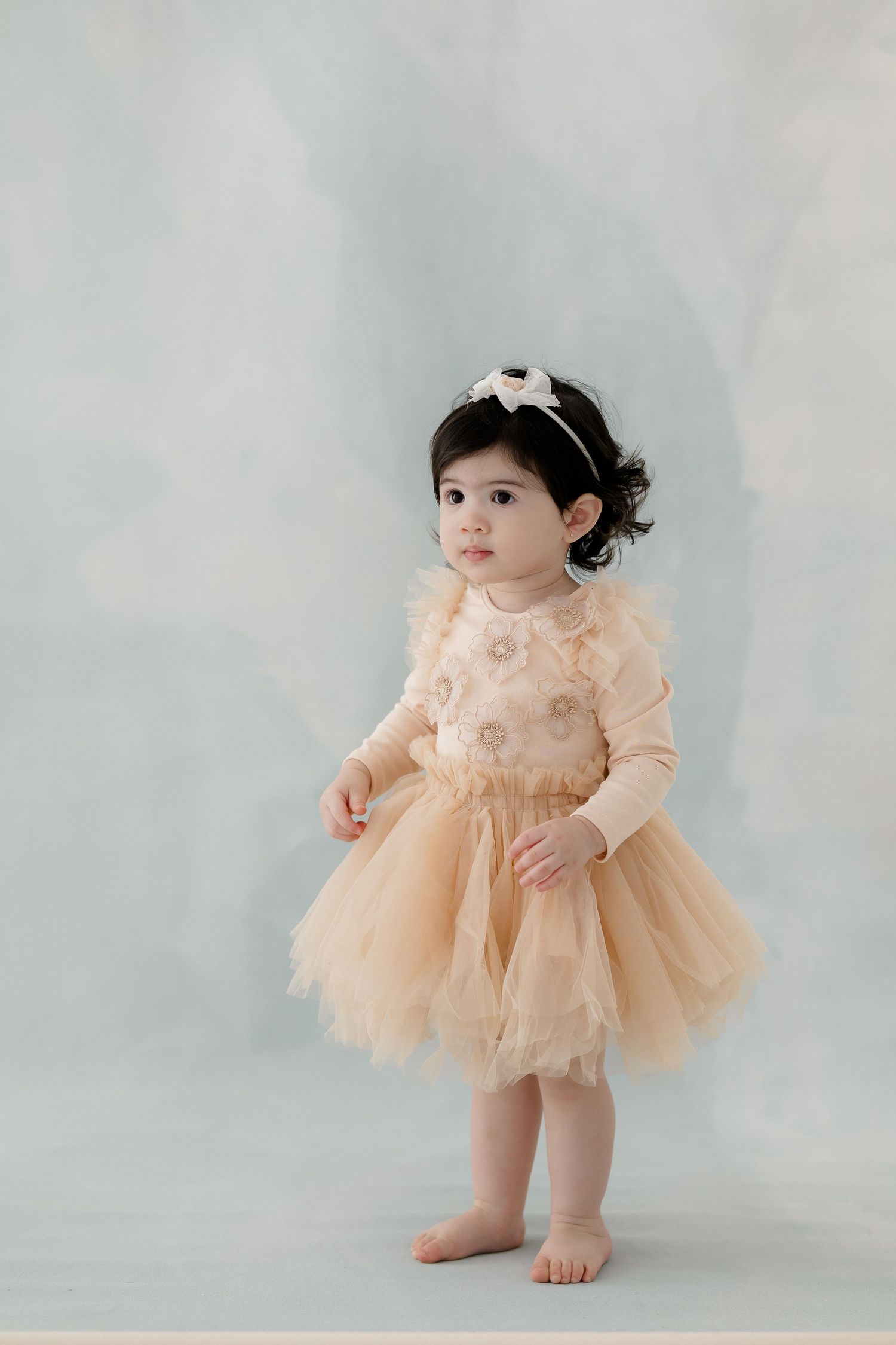 A small child wearing a peach-colored tulle dress and hair bow stands barefoot against a light gray background.