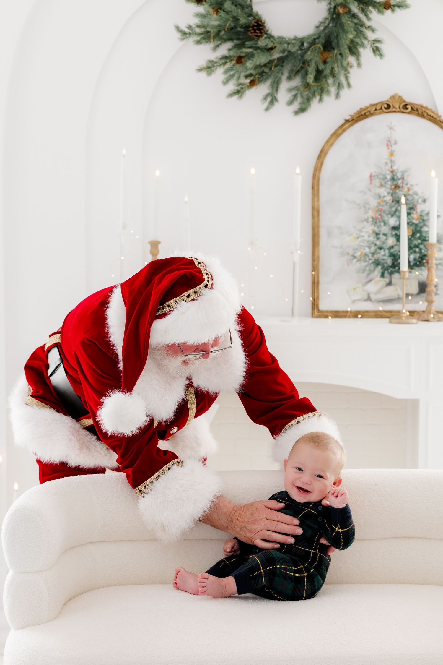 Santa Claus leaning over a smiling baby in plaid outfit on white sofa with Christmas wreath and tree backdrop.