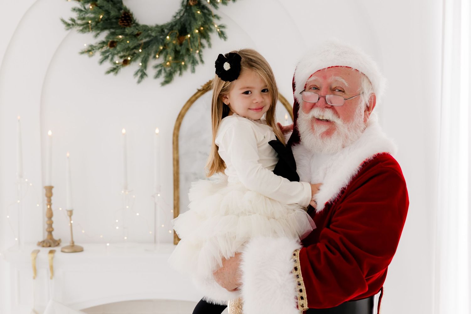 Santa Claus holds a smiling little girl in a white dress during a festive Christmas portrait session.
