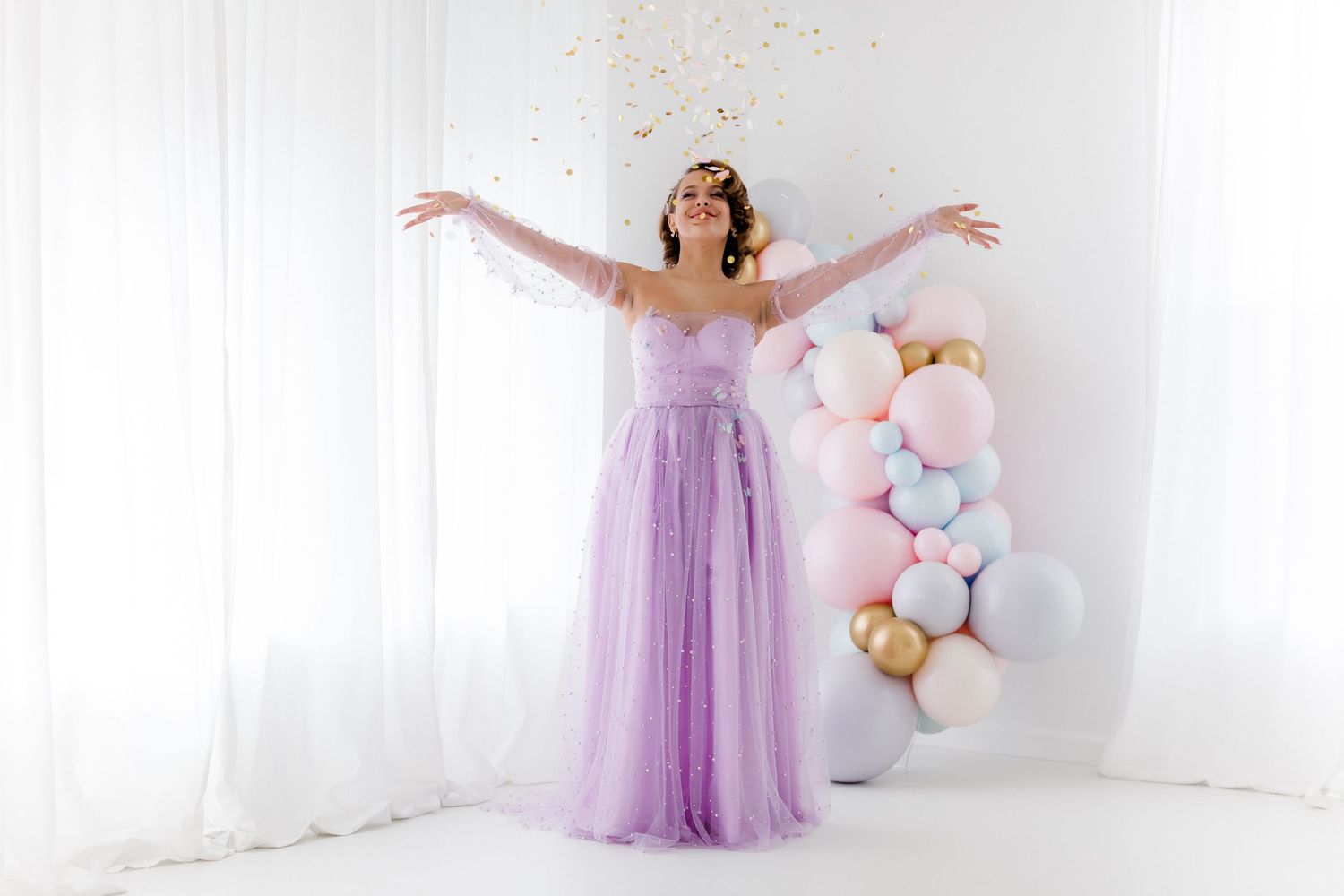 A woman in a flowing lavender gown poses elegantly in a bright white studio with pink balloon decorations.