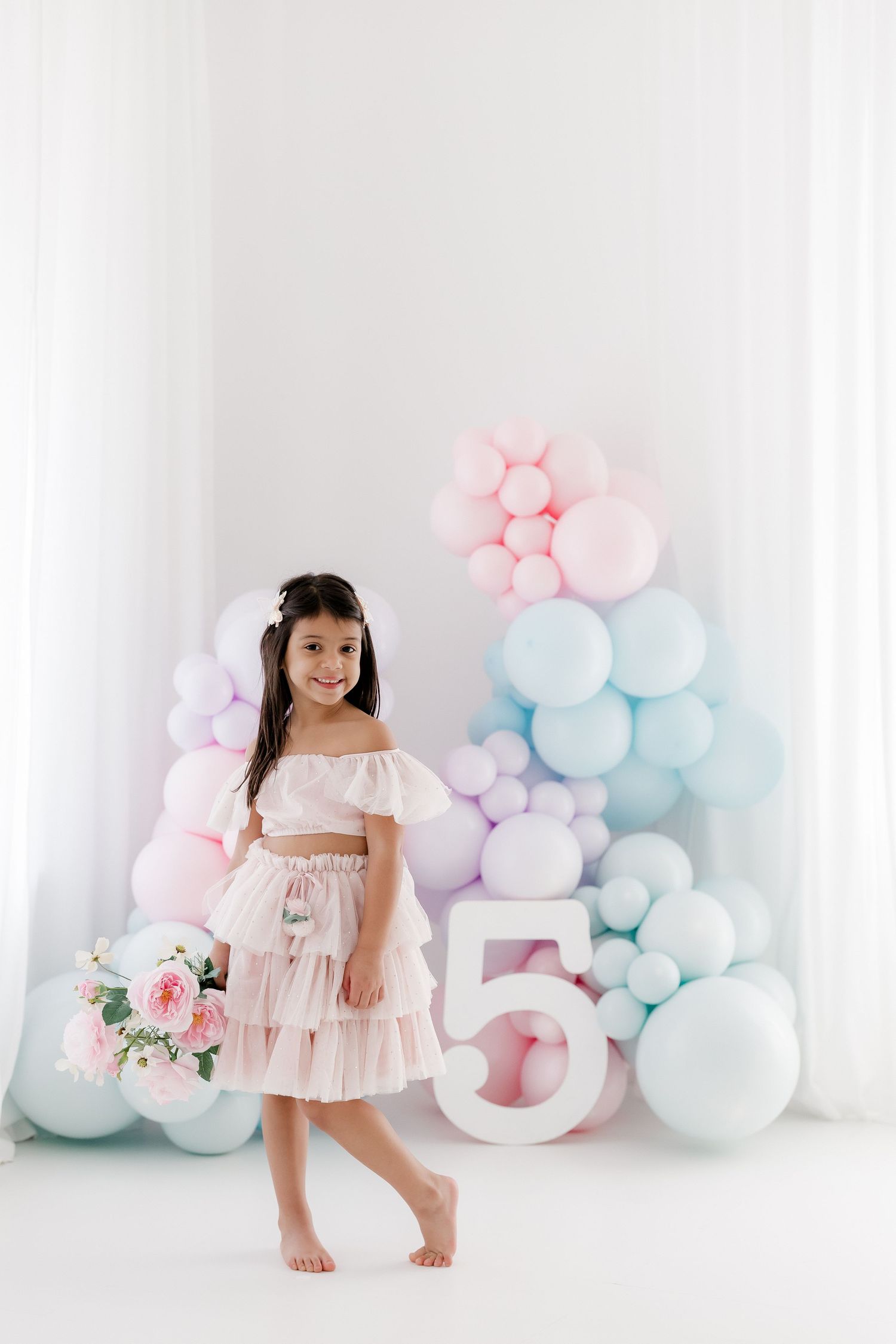 A smiling girl in a pink dress holds flowers beside a large number 5, surrounded by pastel balloons for her birthday.
