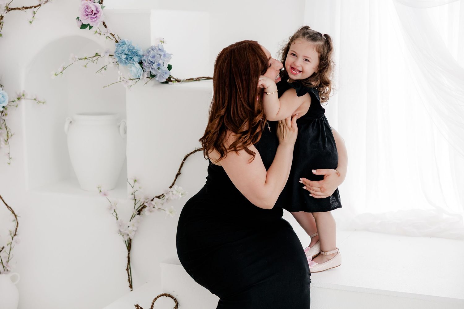 Two people share a tender moment in a bright white studio with delicate floral artwork on the walls.