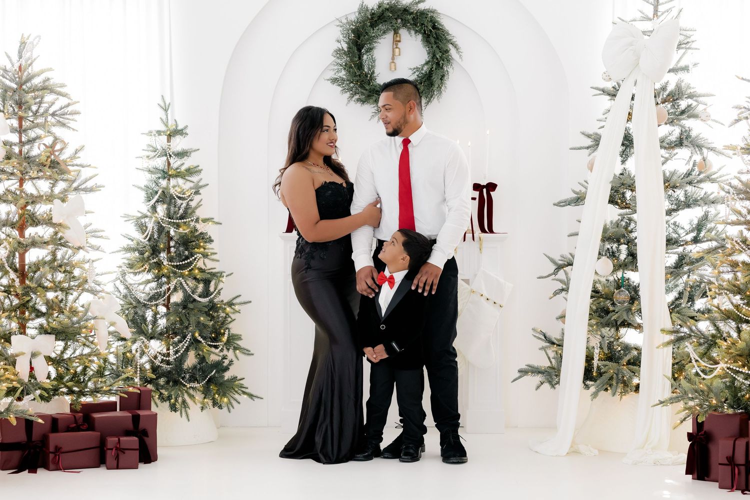 Family of three in elegant holiday attire posing among Christmas trees and gifts in a white studio.