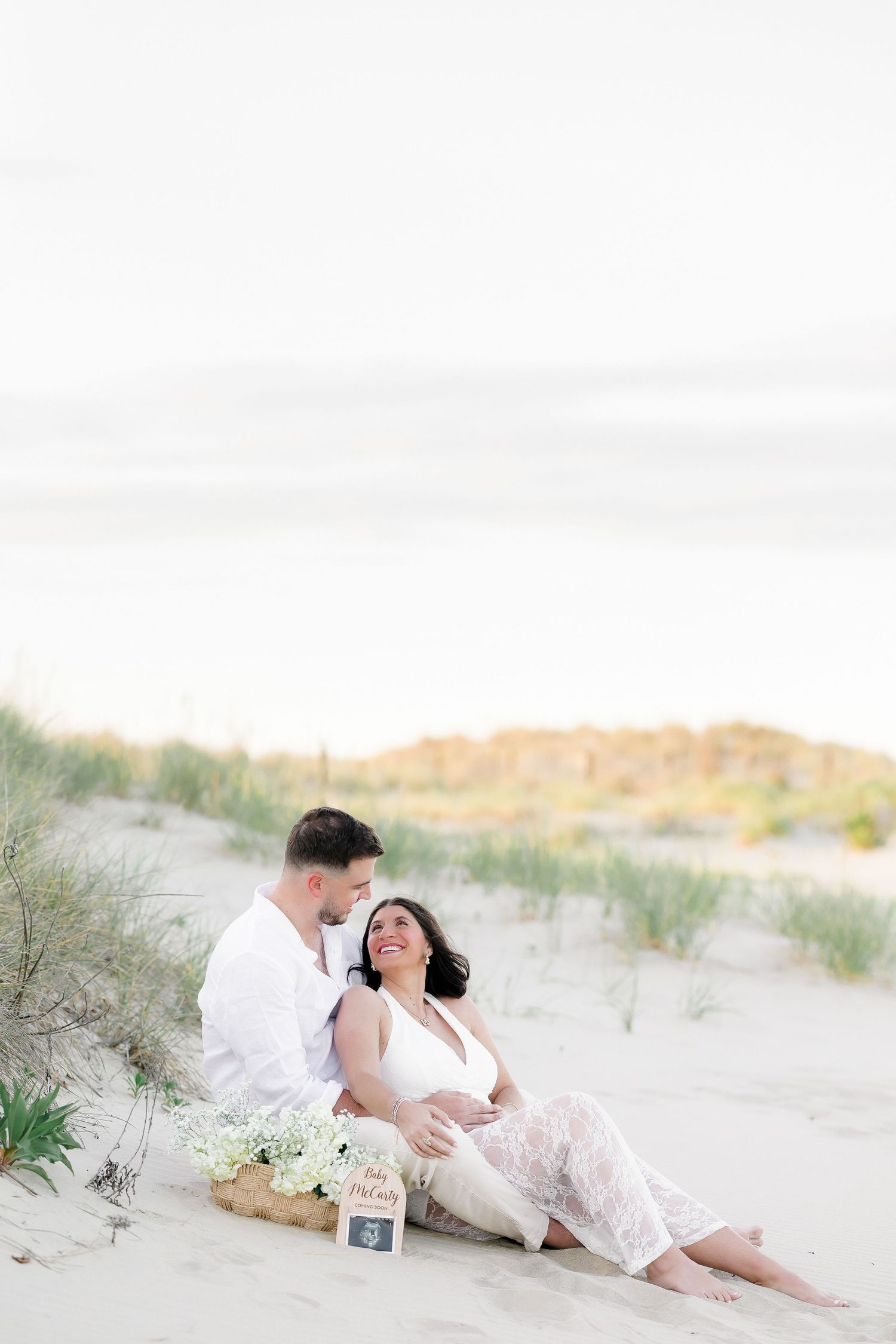 Romantic beach photoshoot featuring couple in white outfits on sandy dunes.