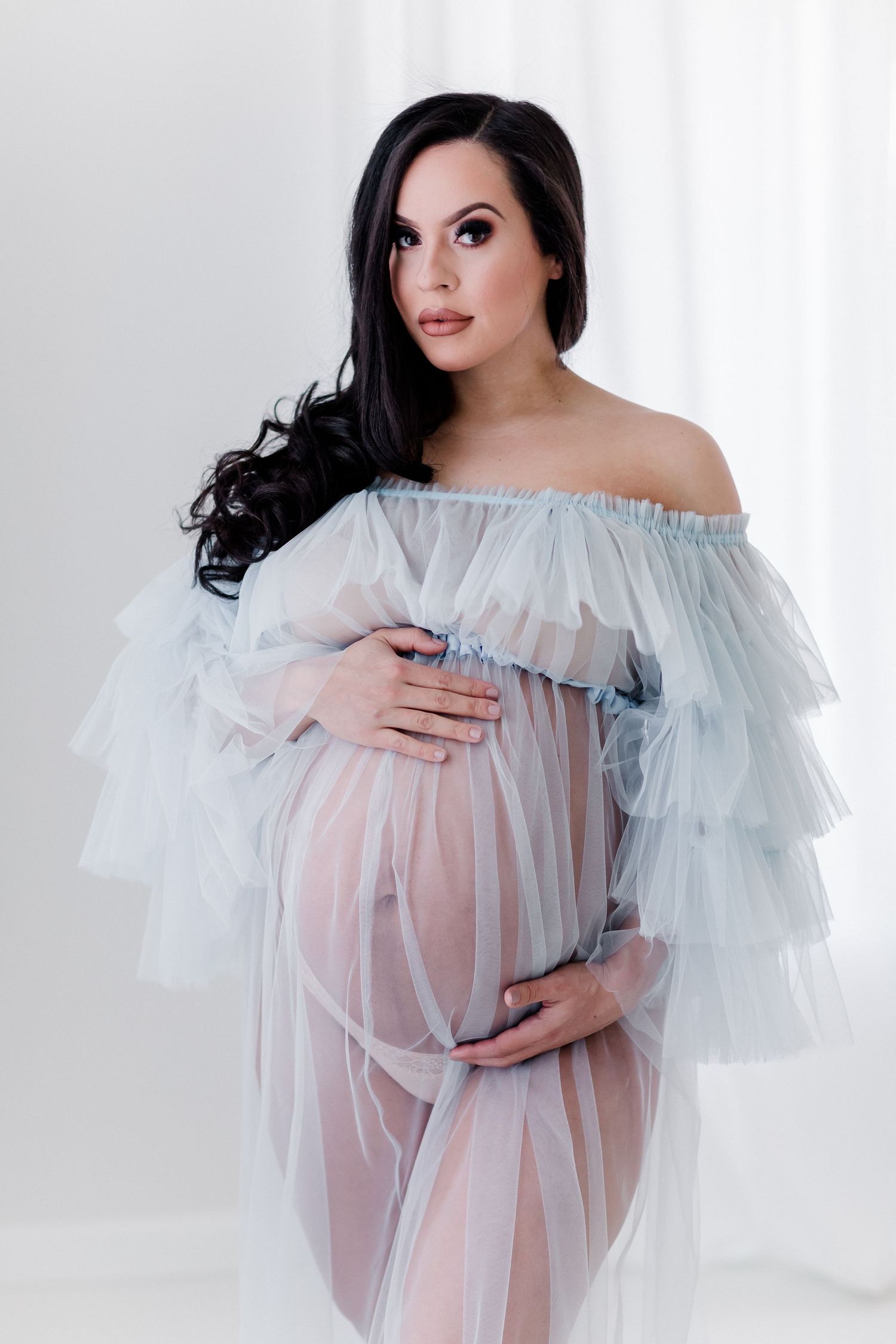 Ethereal maternity photo featuring a sheer off-shoulder tulle gown in a bright white studio setting.