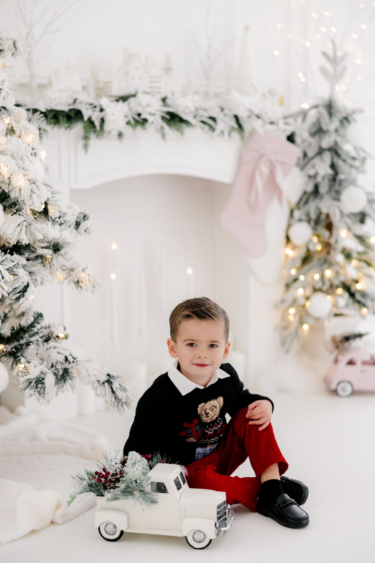 Holiday photoshoot featuring young child with vintage toy car in snowy white studio setting.