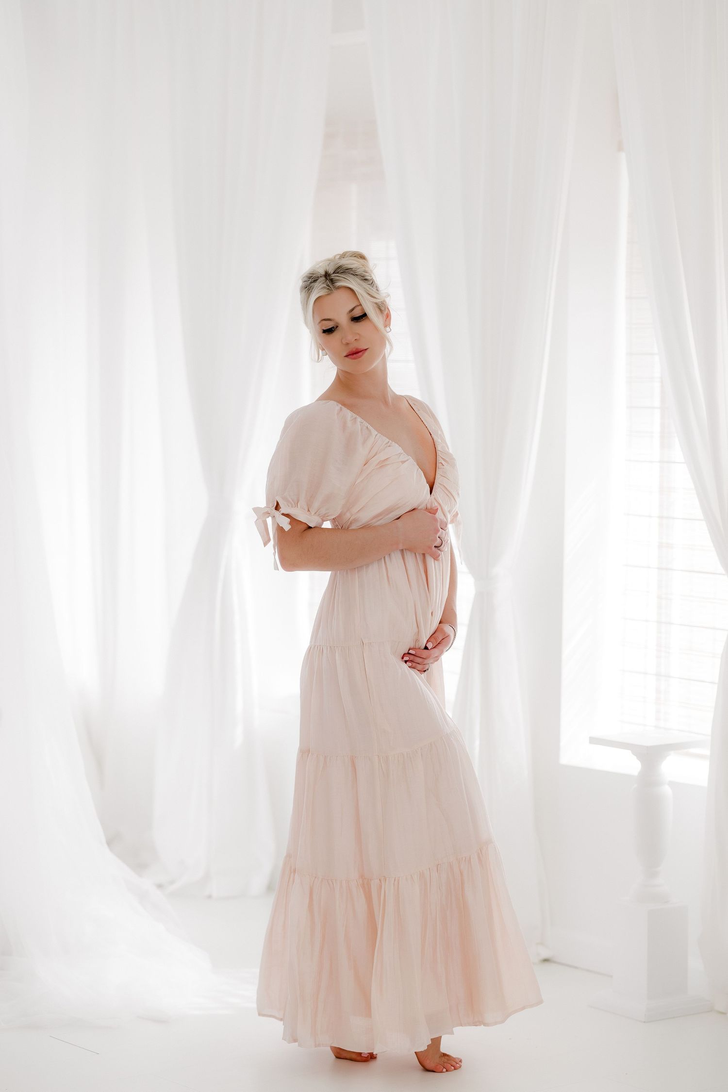 A person wearing a flowing light pink maxi dress poses gracefully against a bright white backdrop with sheer curtains.