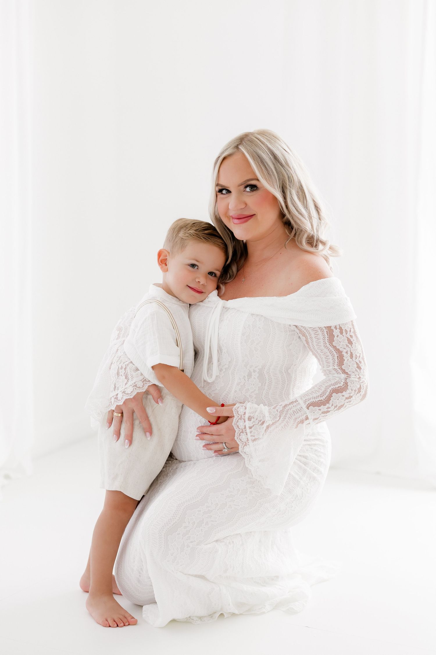 A tender moment captured in white as a mother and child in matching lace dresses embrace against a bright background.