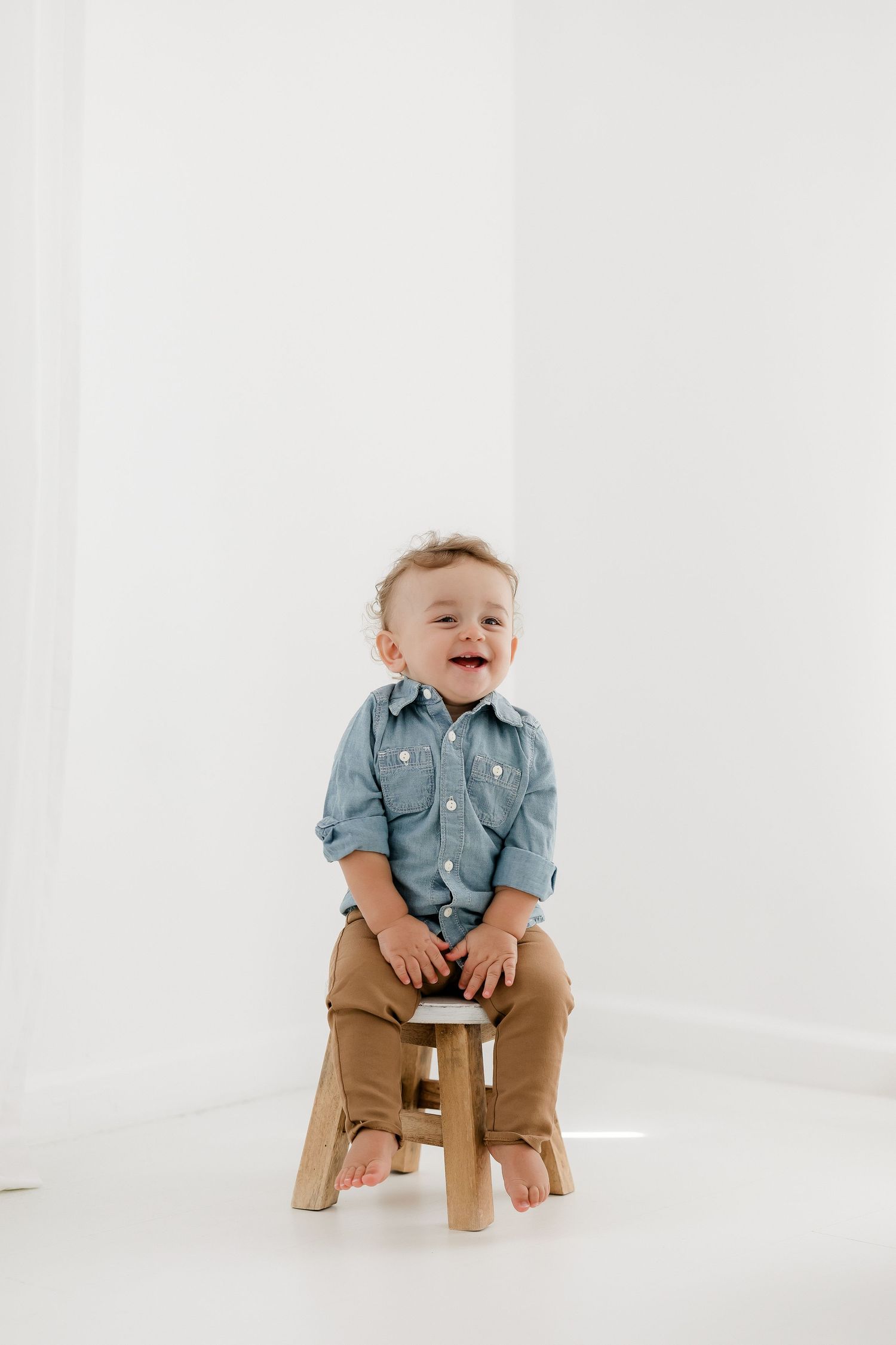 Laughing toddler in chambray shirt and khaki pants sits barefoot on wooden stool against white backdrop.