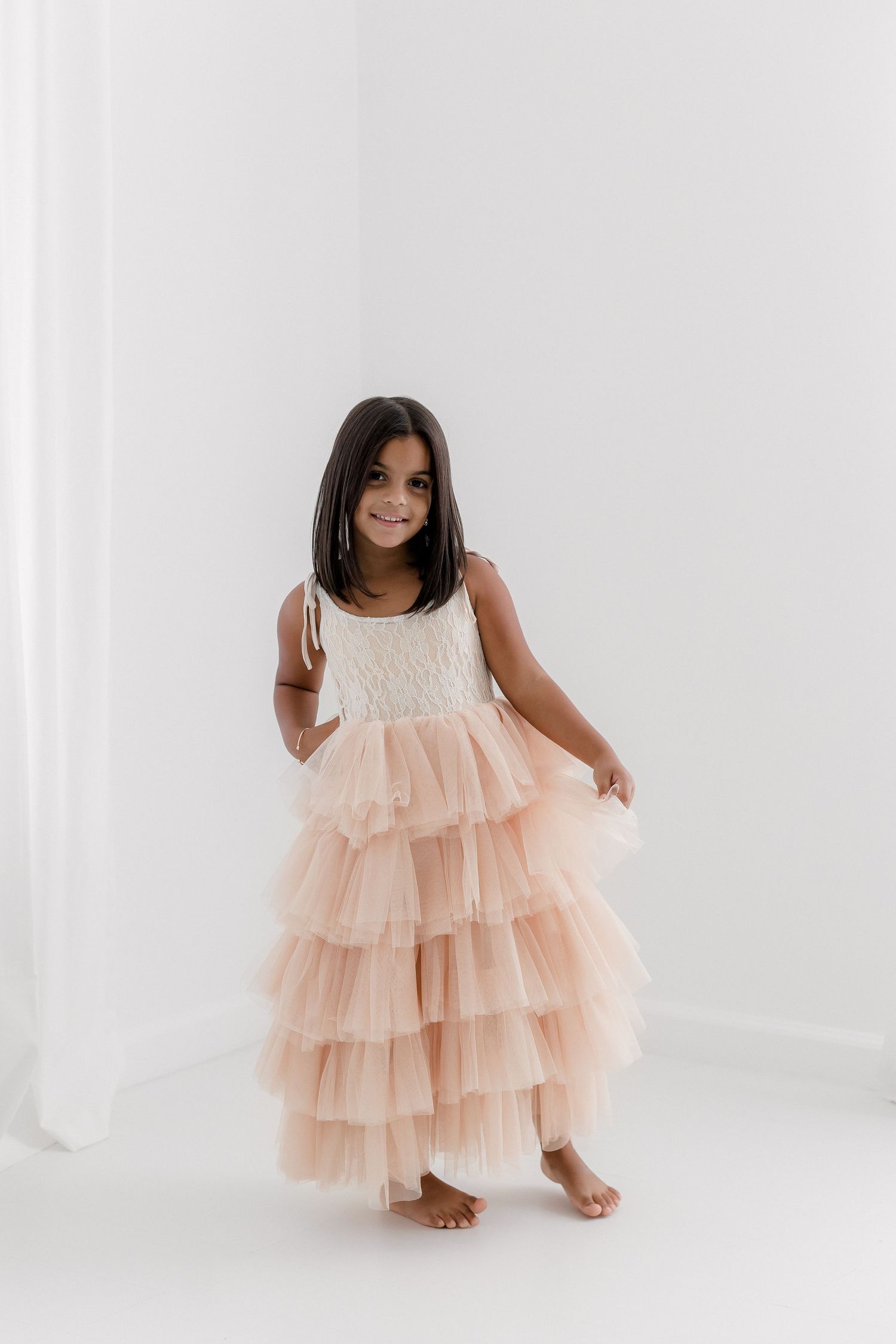 Child wearing tiered peach tulle gown poses gracefully in bright studio.