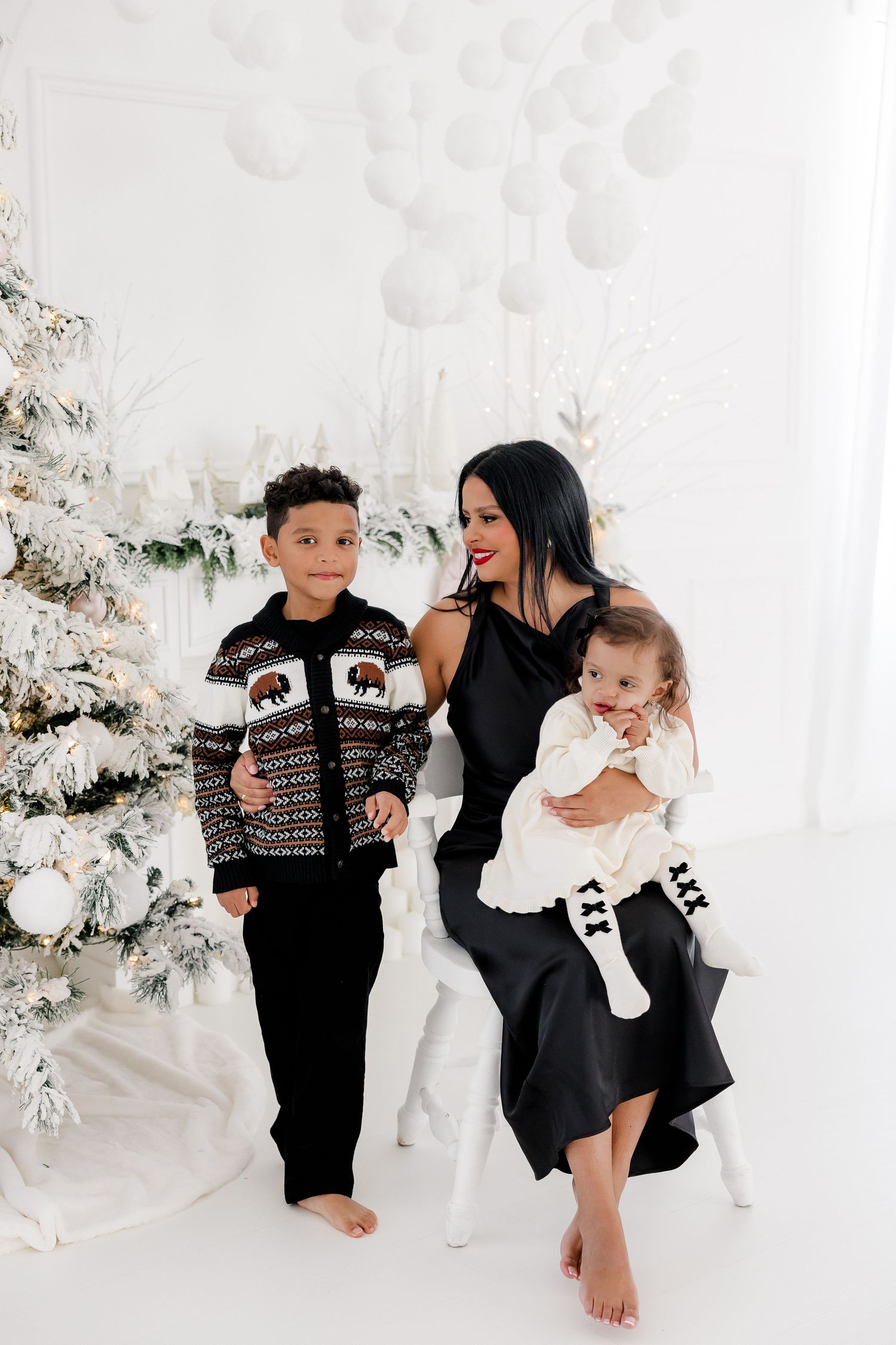 Family sits together in an all-white Christmas studio setting with flocked trees and festive decorations.