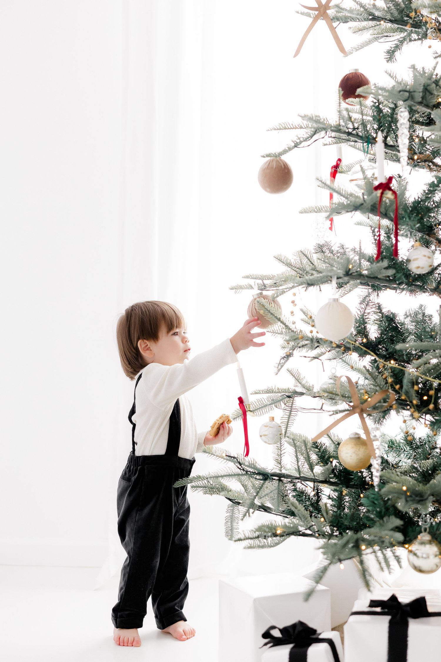 Small child in black and white outfit reaching up to touch ornaments on Christmas tree.