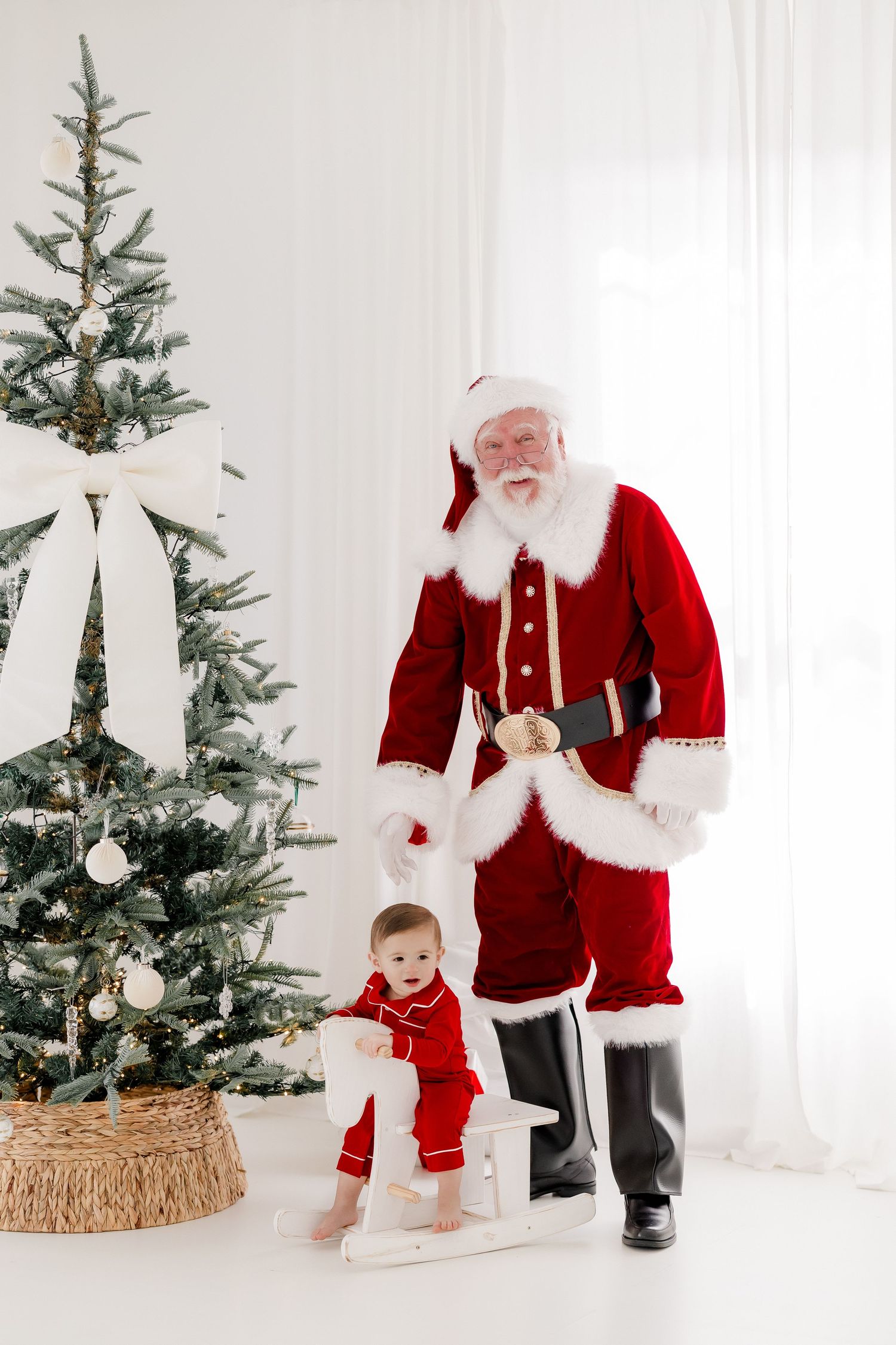 Baby in red pajamas on rocking horse beside Santa Claus and decorated Christmas tree in bright white room.