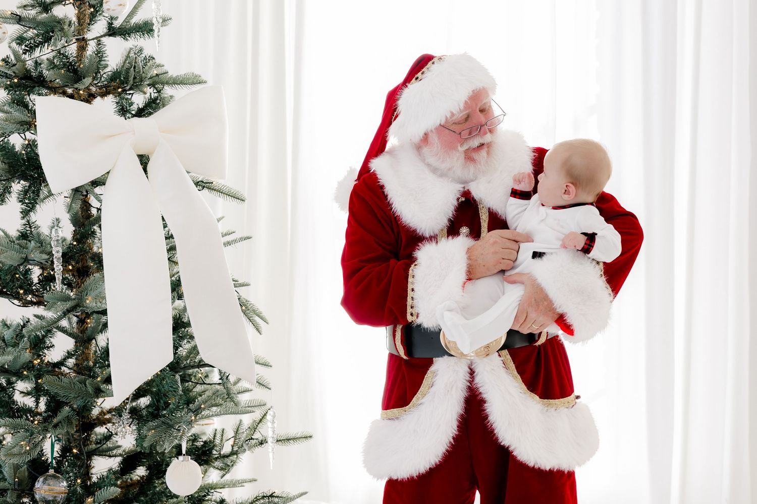 Santa Claus holds a newborn baby in white outfit beside a decorated Christmas tree with white bow.