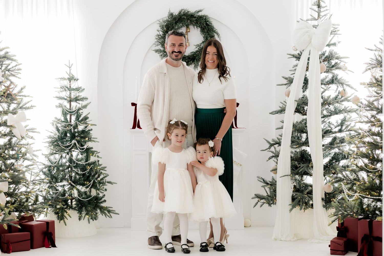 Family of four posing for a Christmas photo with decorated trees and festive wreath backdrop.