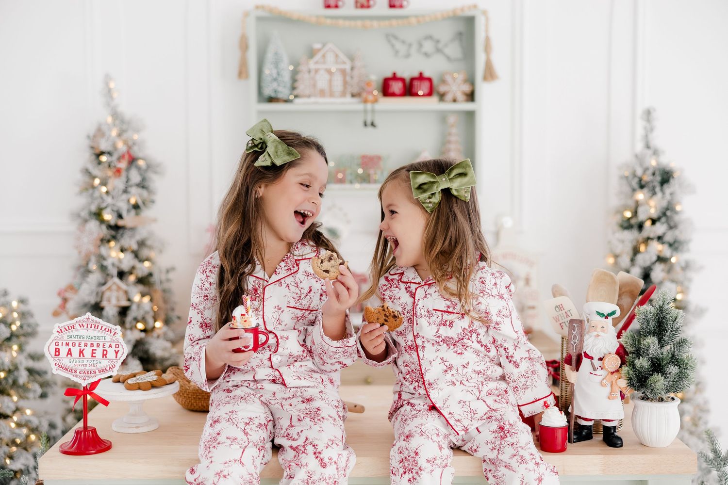 Two girls in matching floral Christmas pajamas eating cookies by a decorated holiday tree.